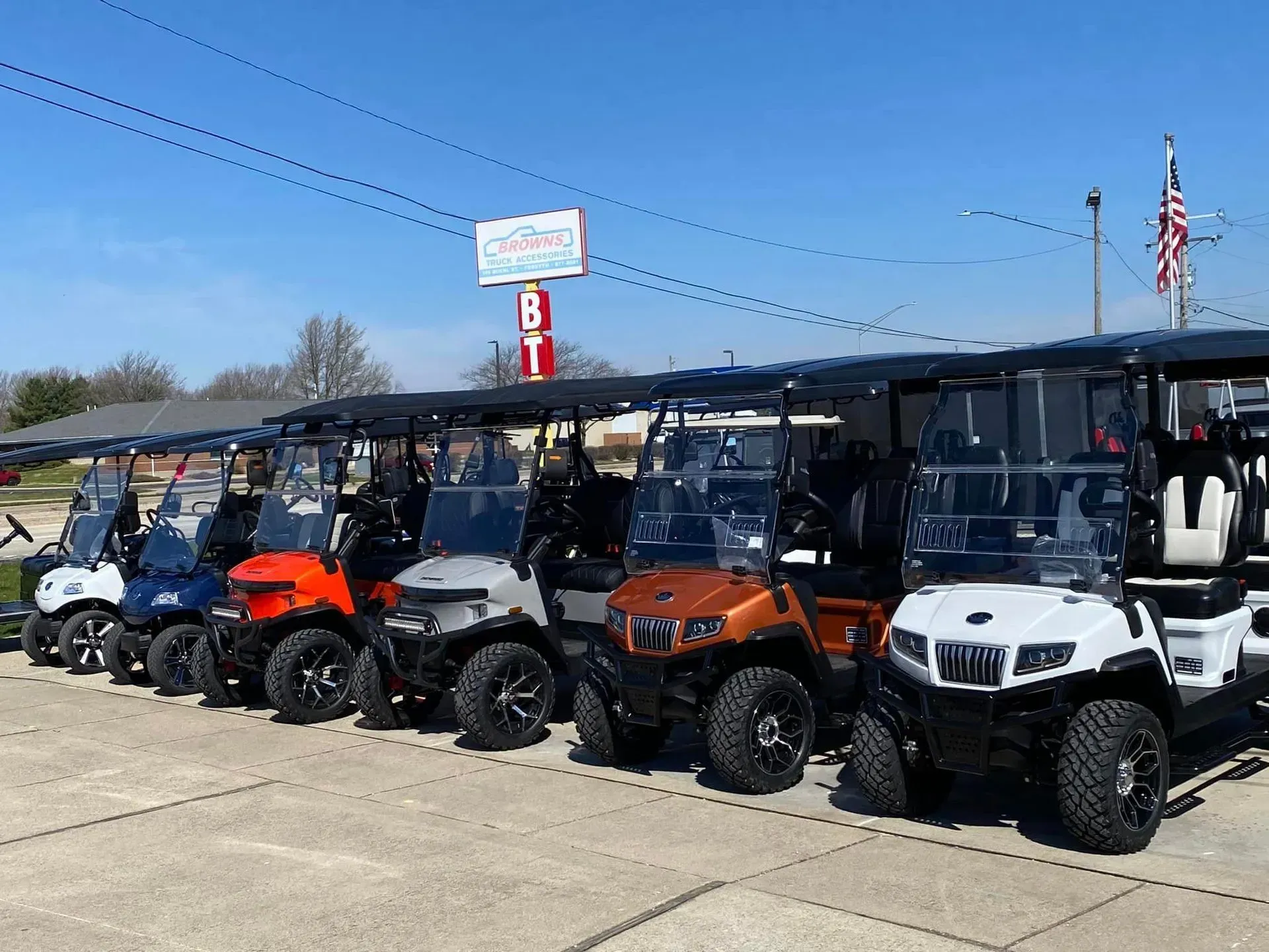 Golf carts in various colors parked in a row, under a canopy at a dealership.