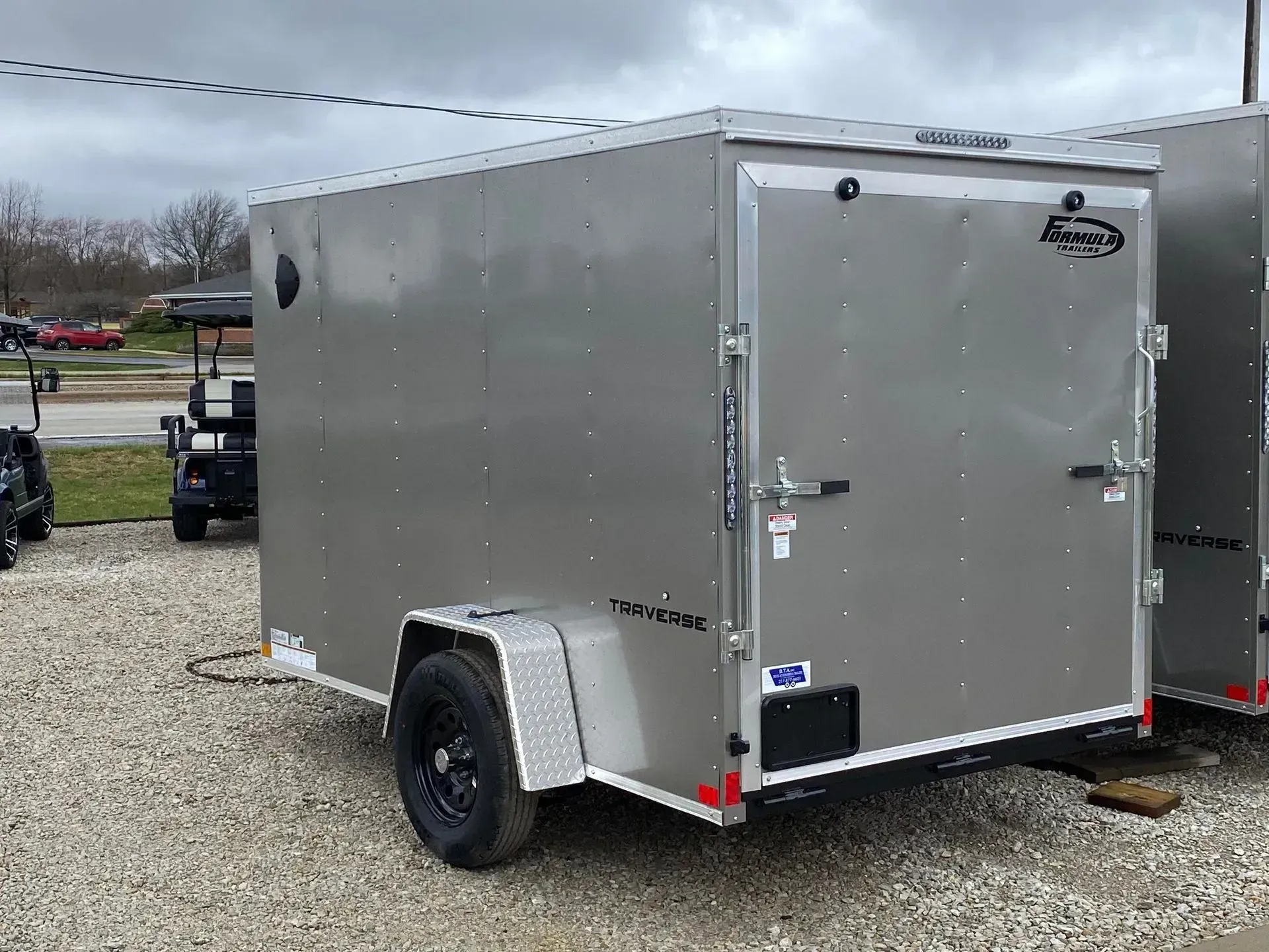 Gray cargo trailer with black wheel and a rear door. Gravel ground.