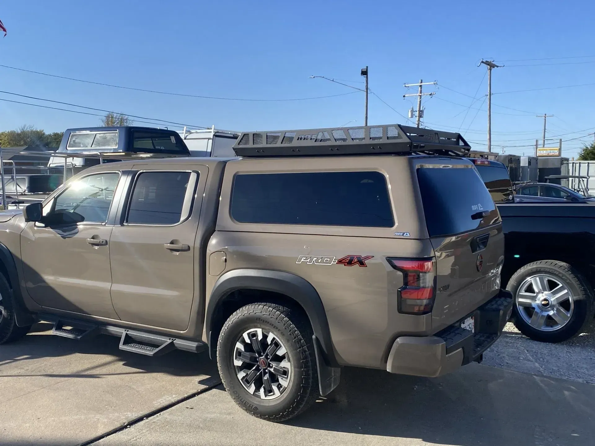 Tan Nissan Frontier truck with a camper shell and roof rack, parked outside on a sunny day.