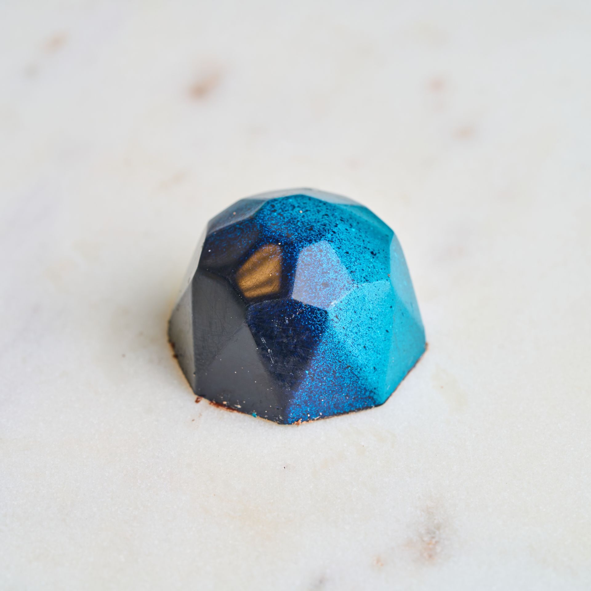 Blue and black faceted chocolate candy on a marble surface.