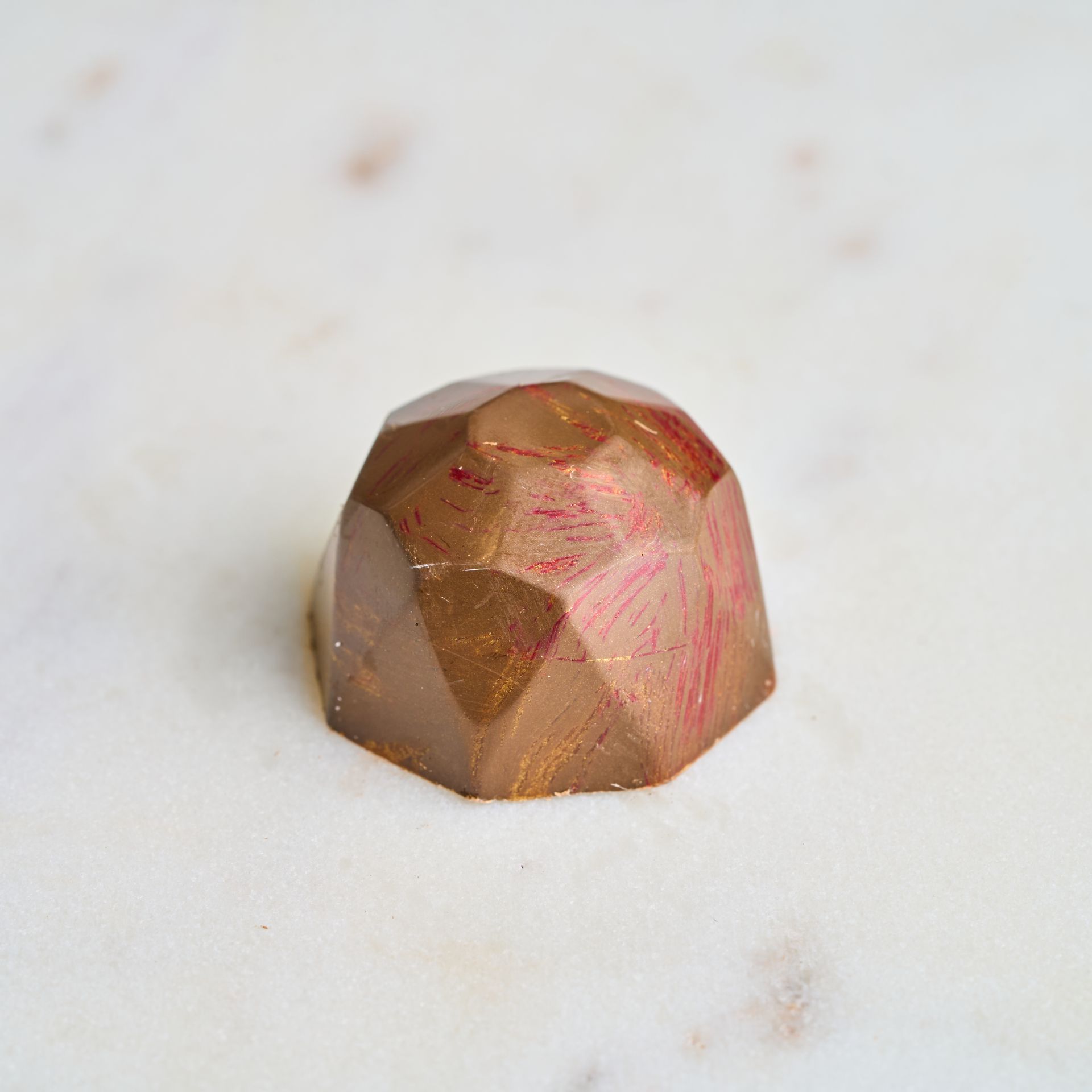 A faceted, jewel-shaped chocolate truffle with gold and red hues on a marble surface.