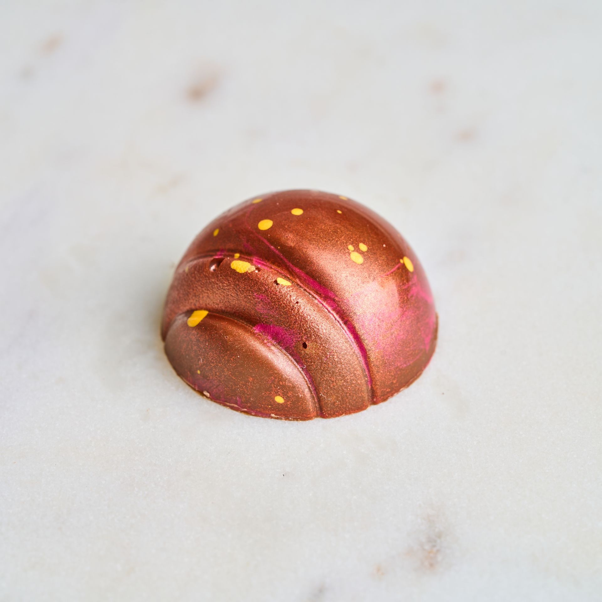 Chocolate truffle, copper-colored with pink accents and yellow speckles, on a marble surface.