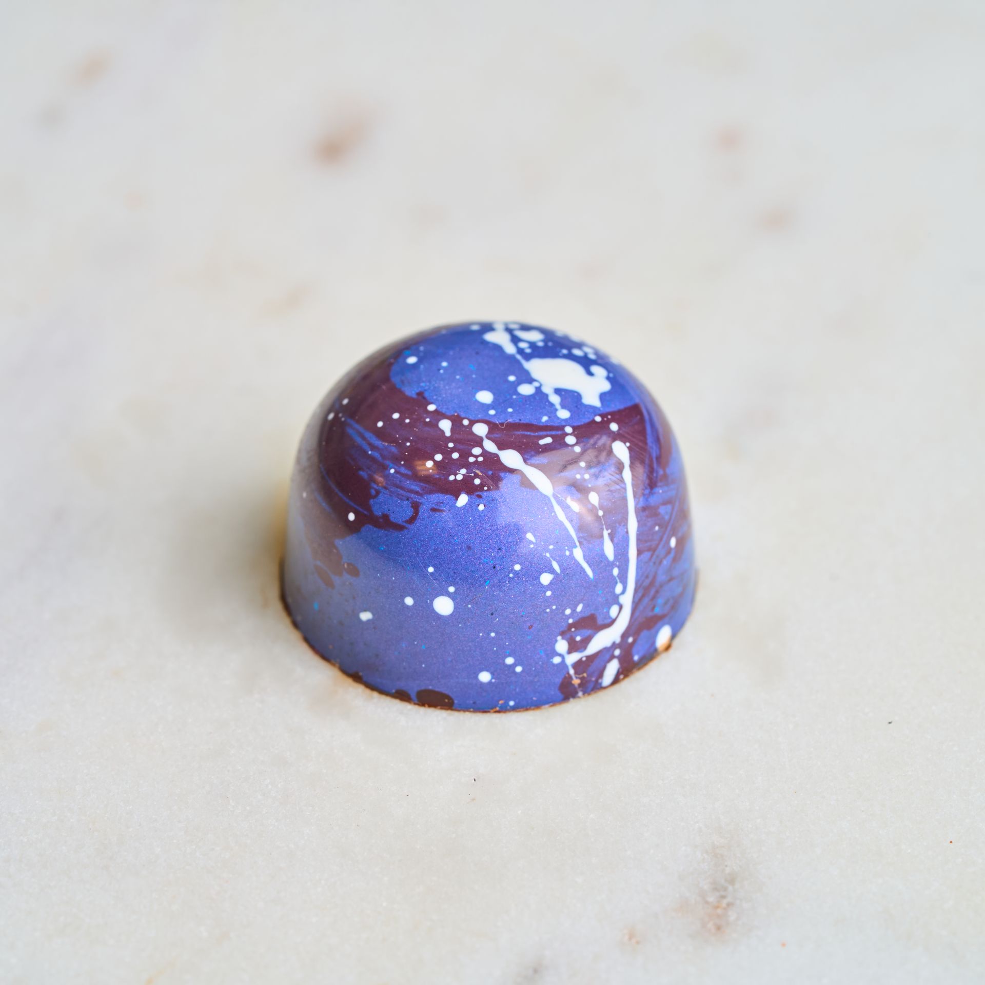 A purple and white speckled chocolate truffle on a white marble surface.