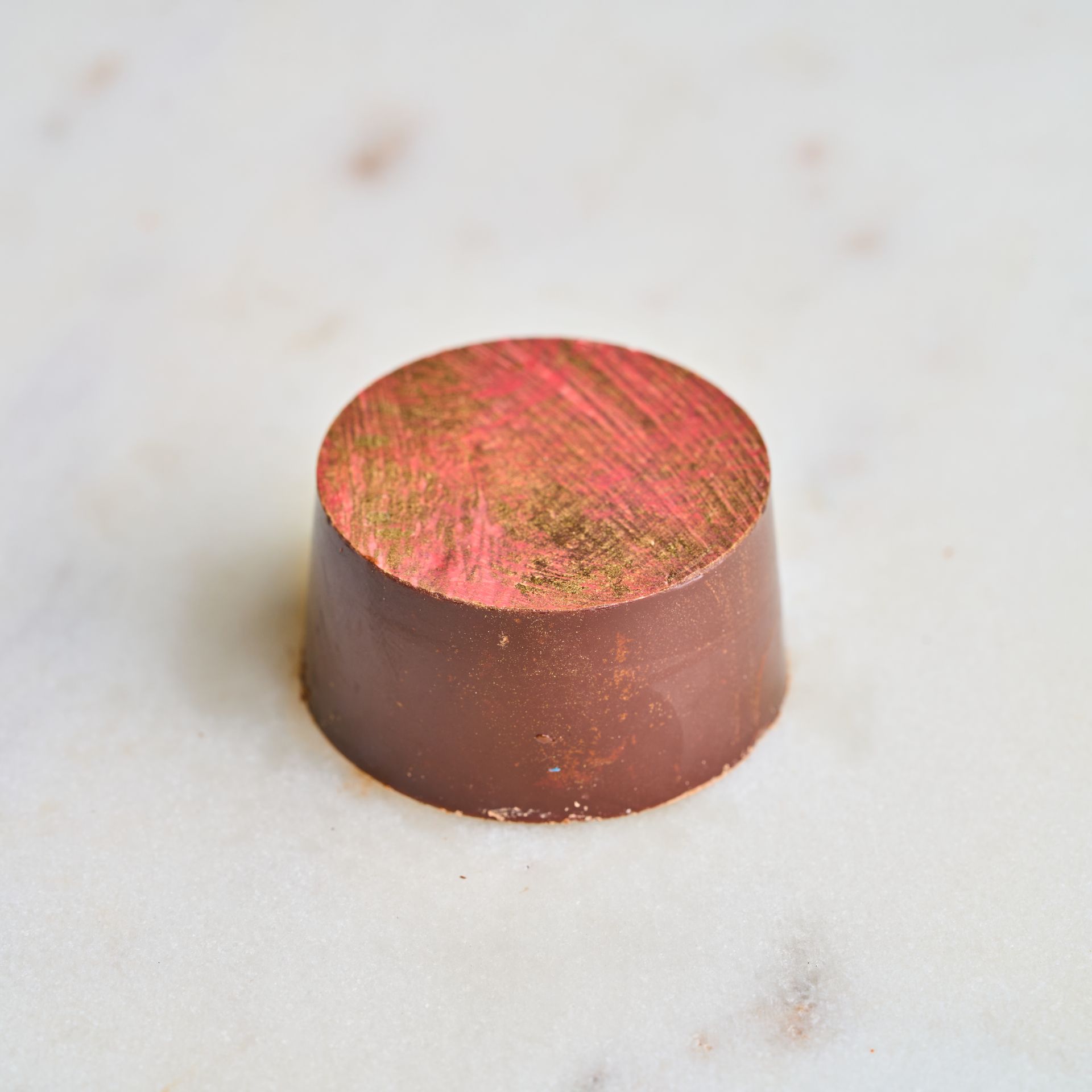 Chocolate candy, cylindrical, brown sides, pink and gold brushed top, on a white surface.