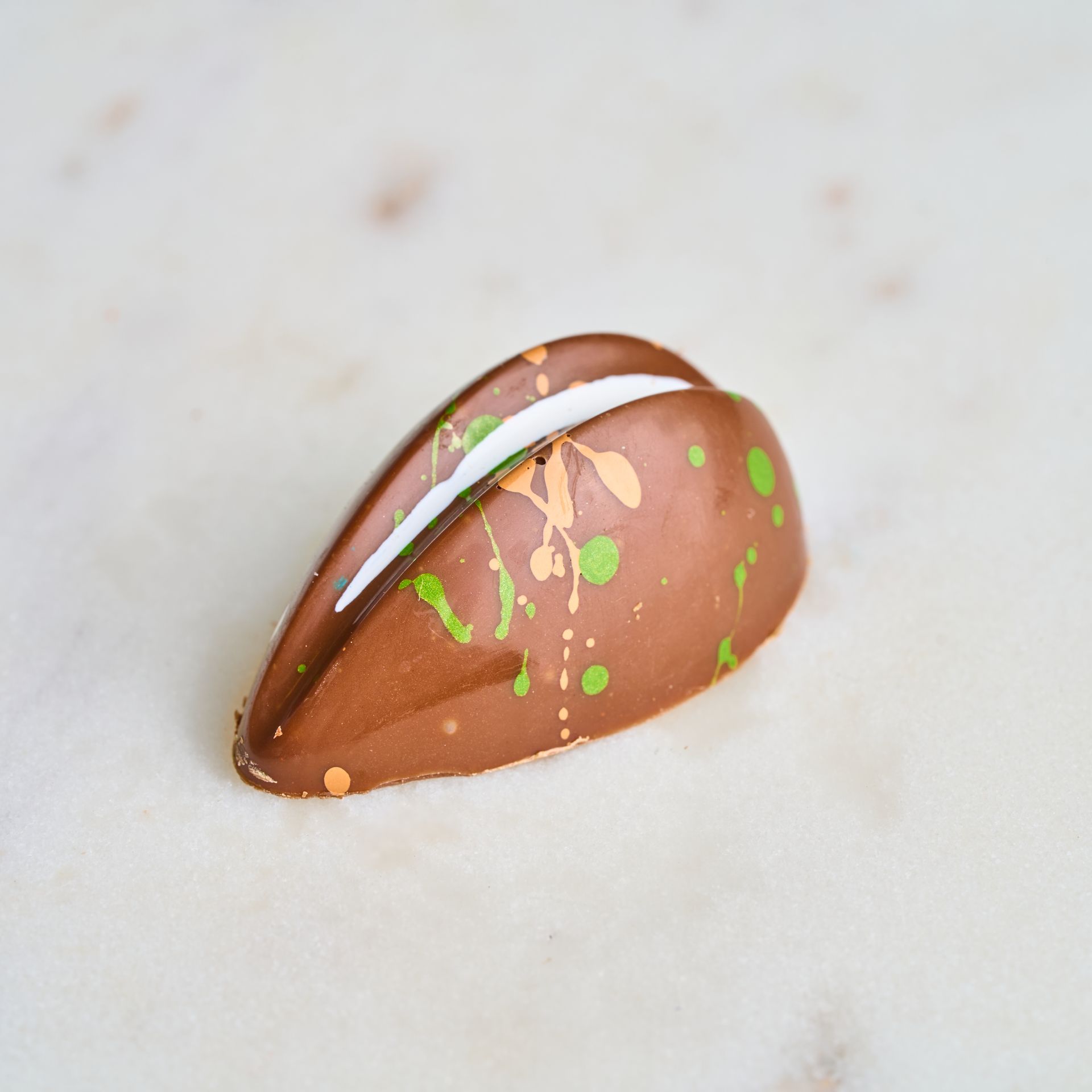 Chocolate candy with white filling, brown coating, and green/orange splatter on a marble surface.