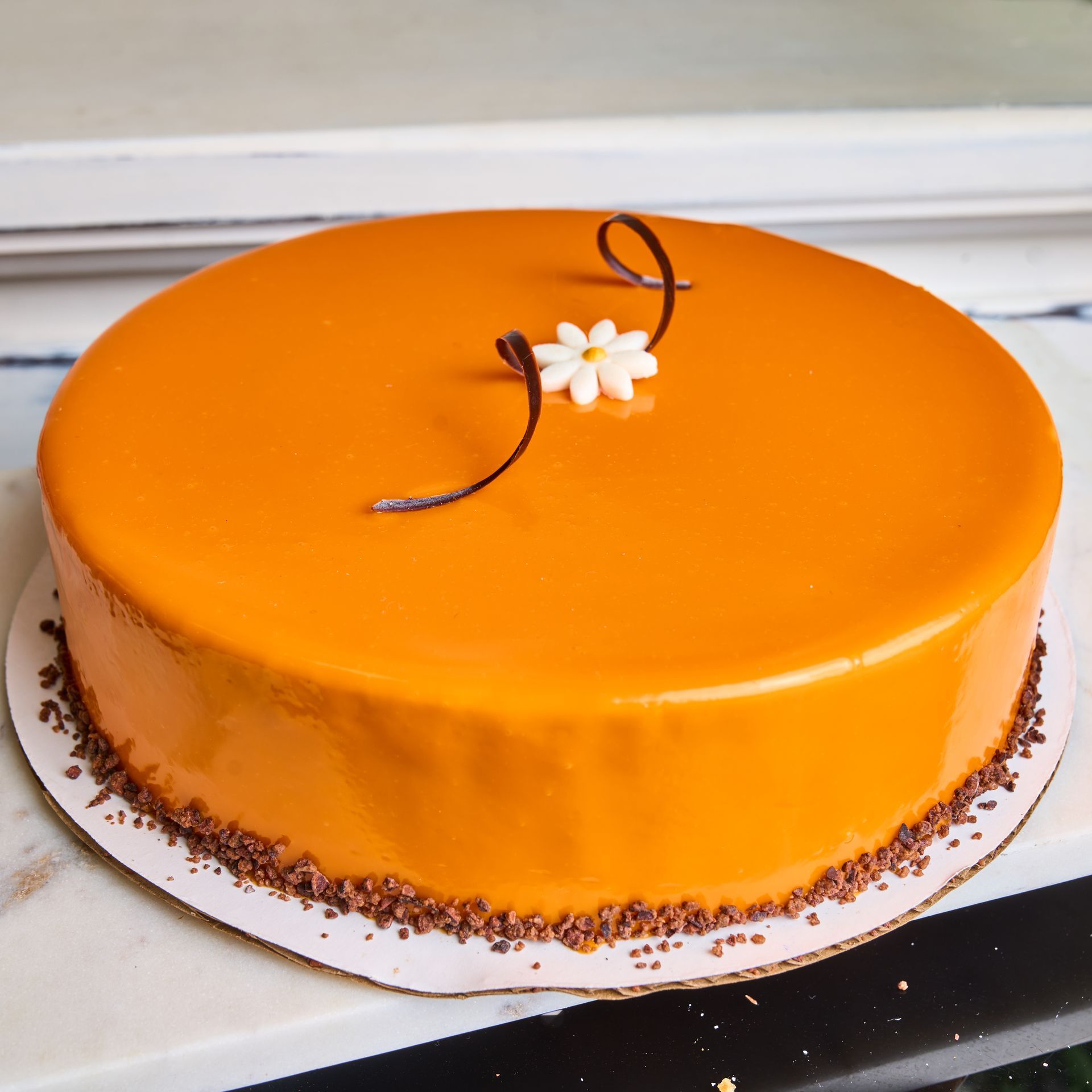 Round orange glazed cake with flower and chocolate curls on a white surface, cake is on a cardboard base.
