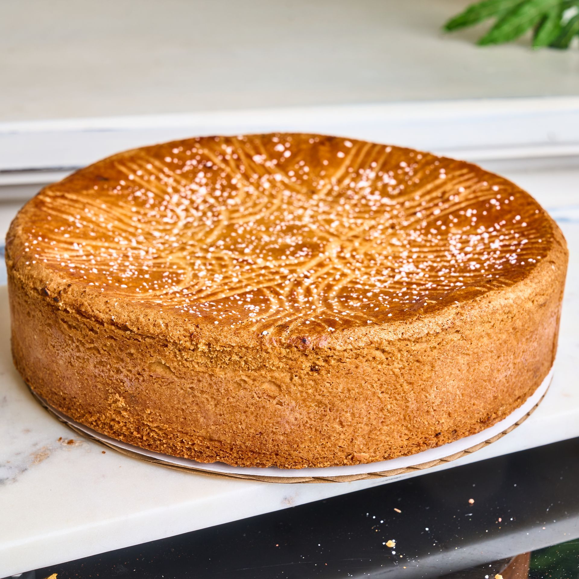 Round golden cake with patterned top, dusted with powdered sugar, on a white surface.