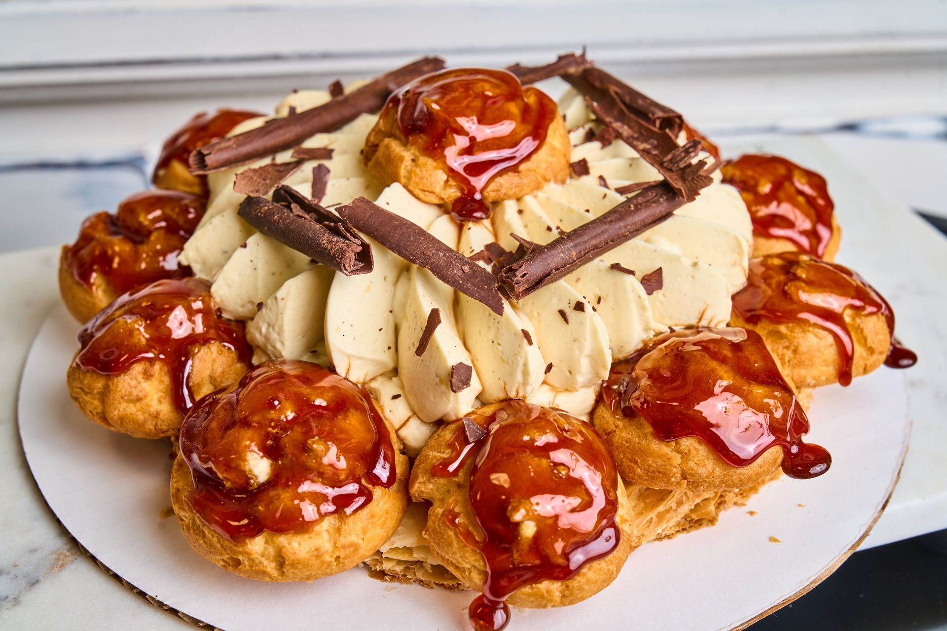 Profiterole cake with caramel sauce, whipped cream, and chocolate decorations on a white plate.