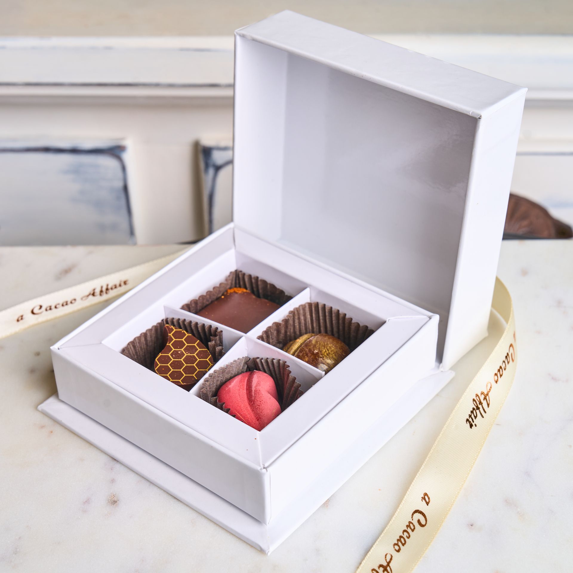 White box of chocolates with four different pieces, ribbon in a white setting.