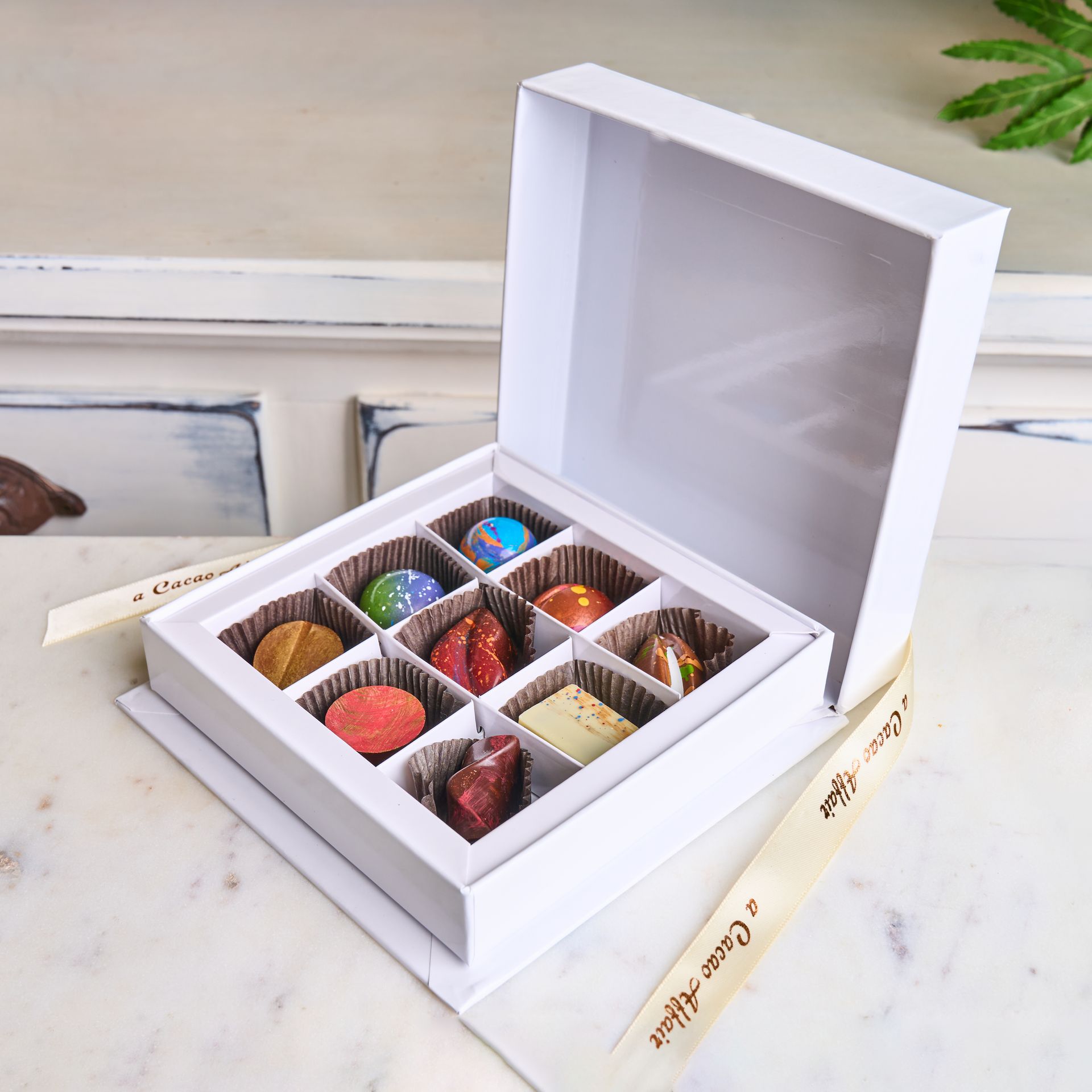 White box of chocolates, lid open, revealing a selection of colorful artisan confections on a white surface.