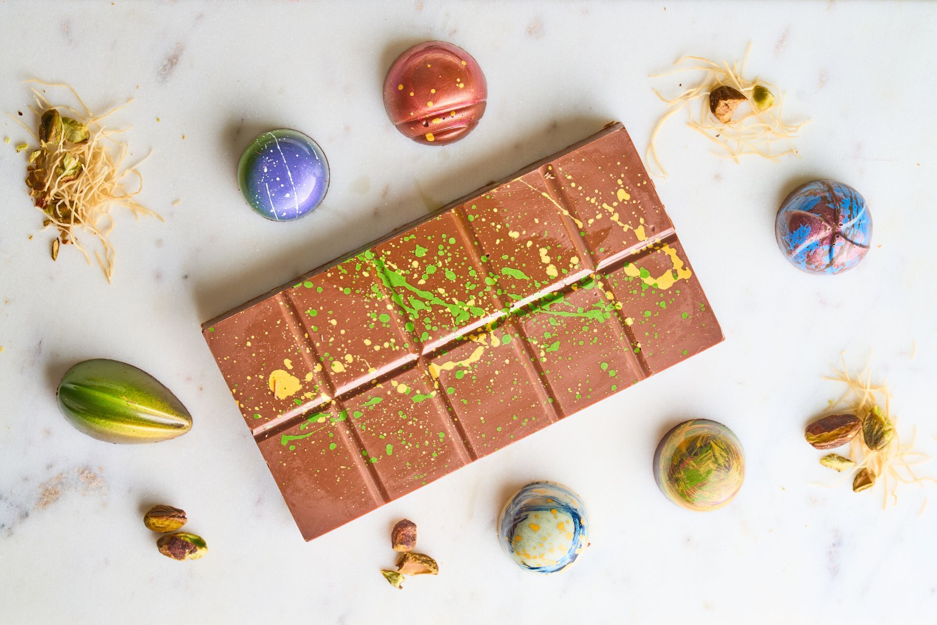 Chocolate bar and truffles on white surface, with scattered pistachios and gold/green accents.