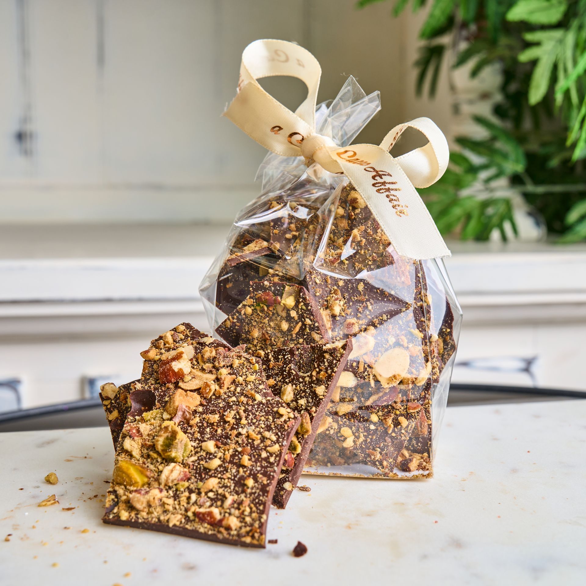 Bag of chocolate bark topped with nuts, tied with a ribbon. Several pieces rest in front.
