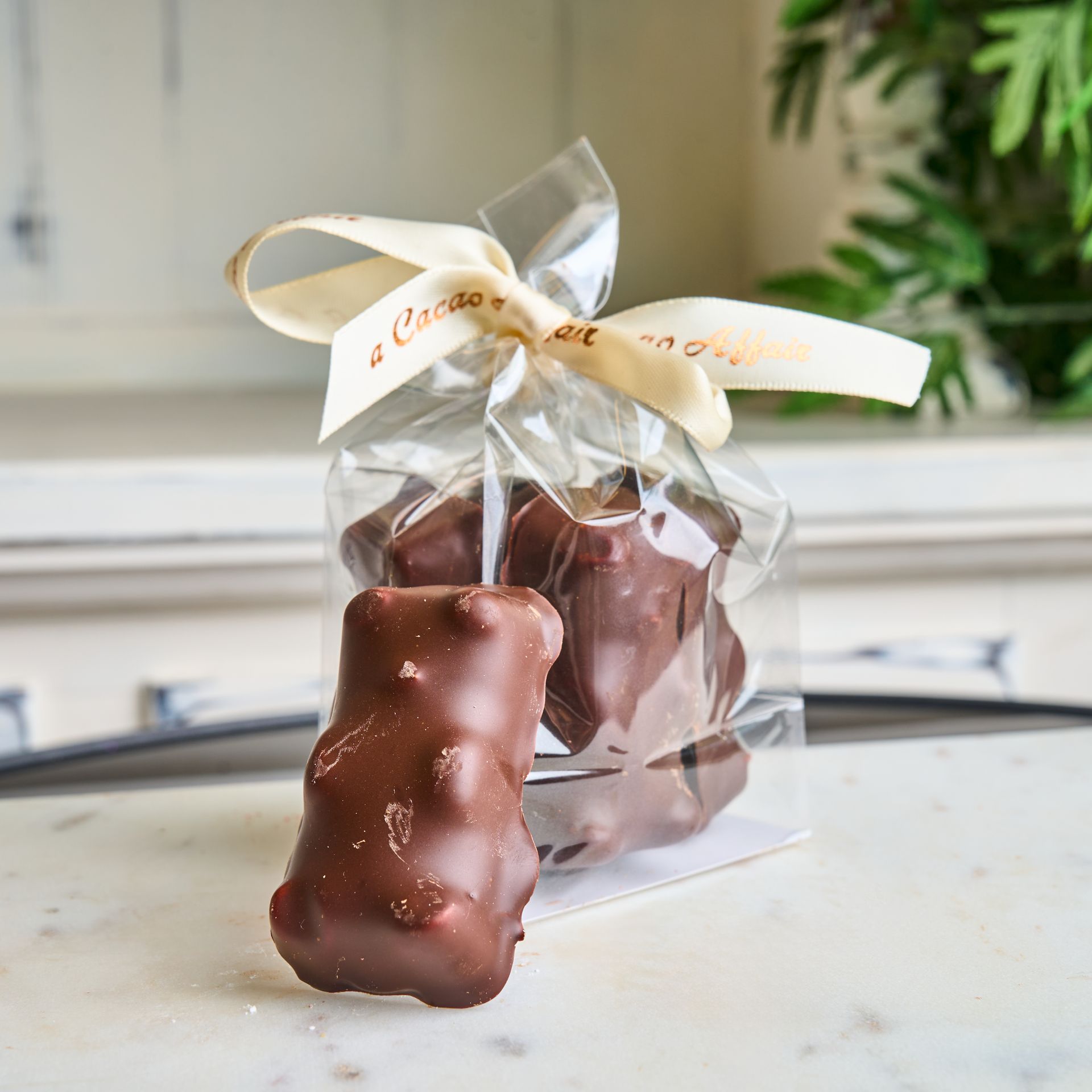 Chocolate-covered gummy bears in a clear bag tied with a cream ribbon on a white marble surface.