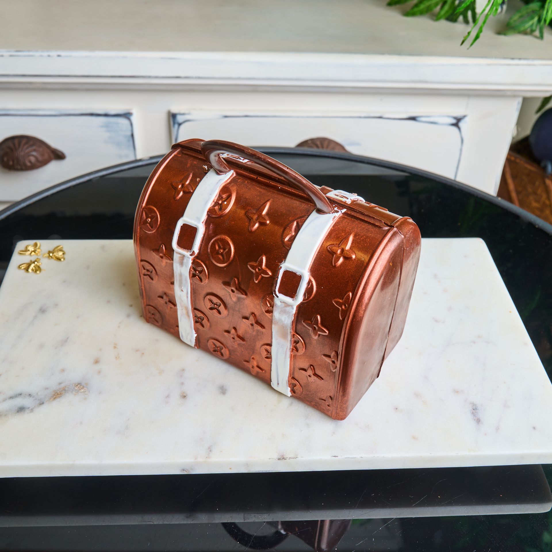 Brown cake shaped like a travel bag, on a marble slab. White straps, gold accents.