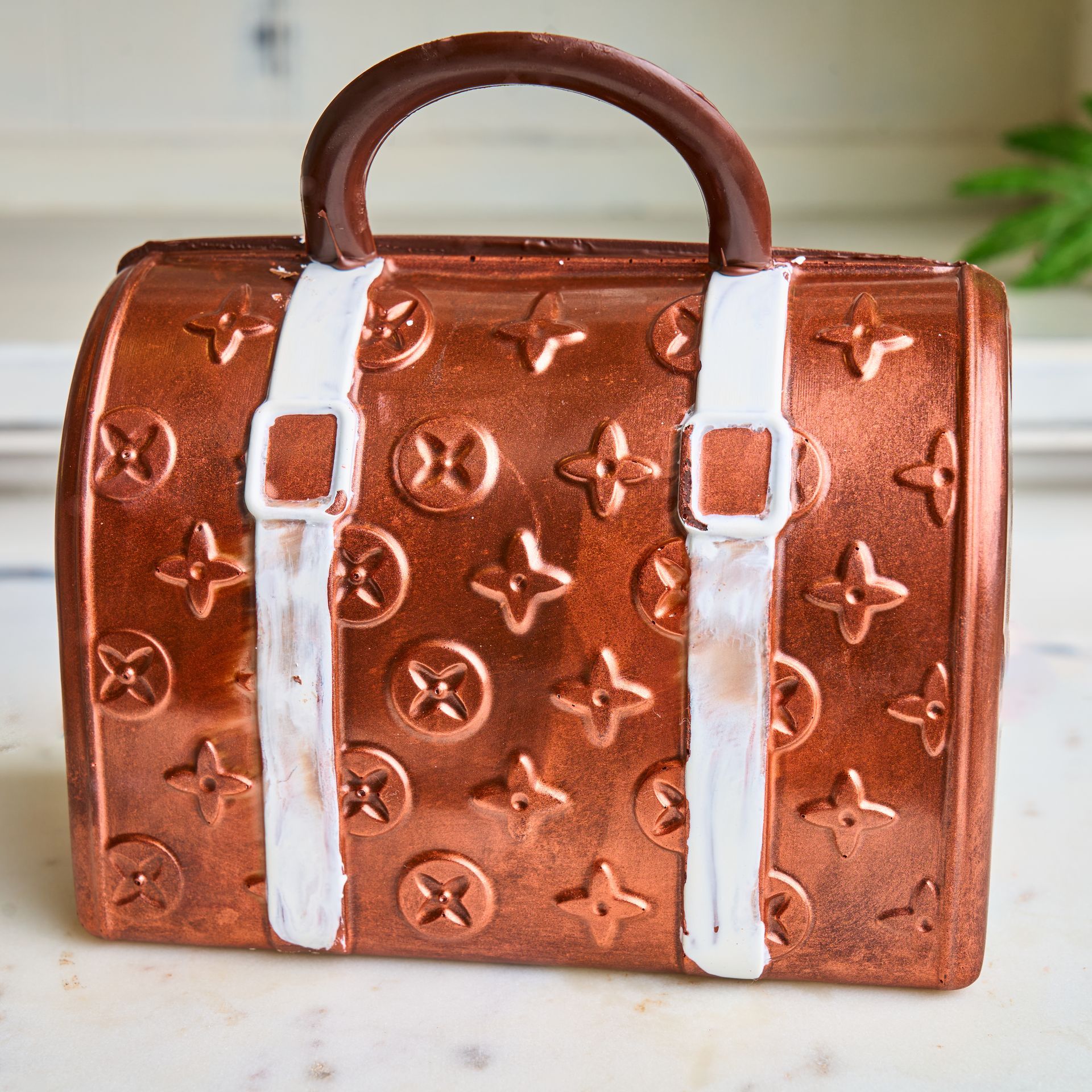 Chocolate purse-shaped confection with brown body, white straps, and brown handle. Decorative logo design visible.