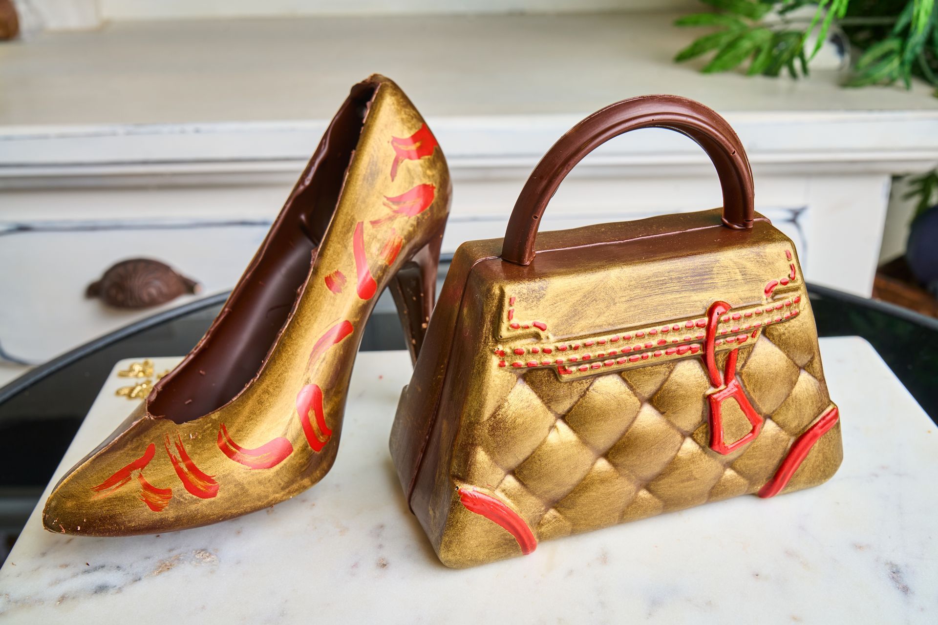 Gold-colored chocolate high heel and handbag on marble surface, with red accents.