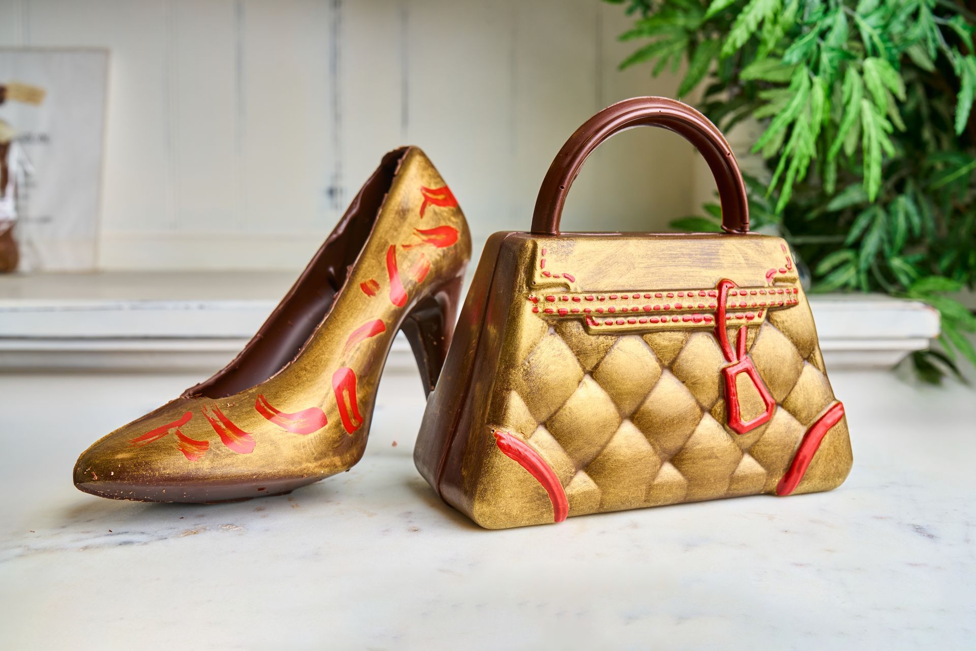 Chocolate high-heeled shoe and handbag, gold with red accents, on a white surface.