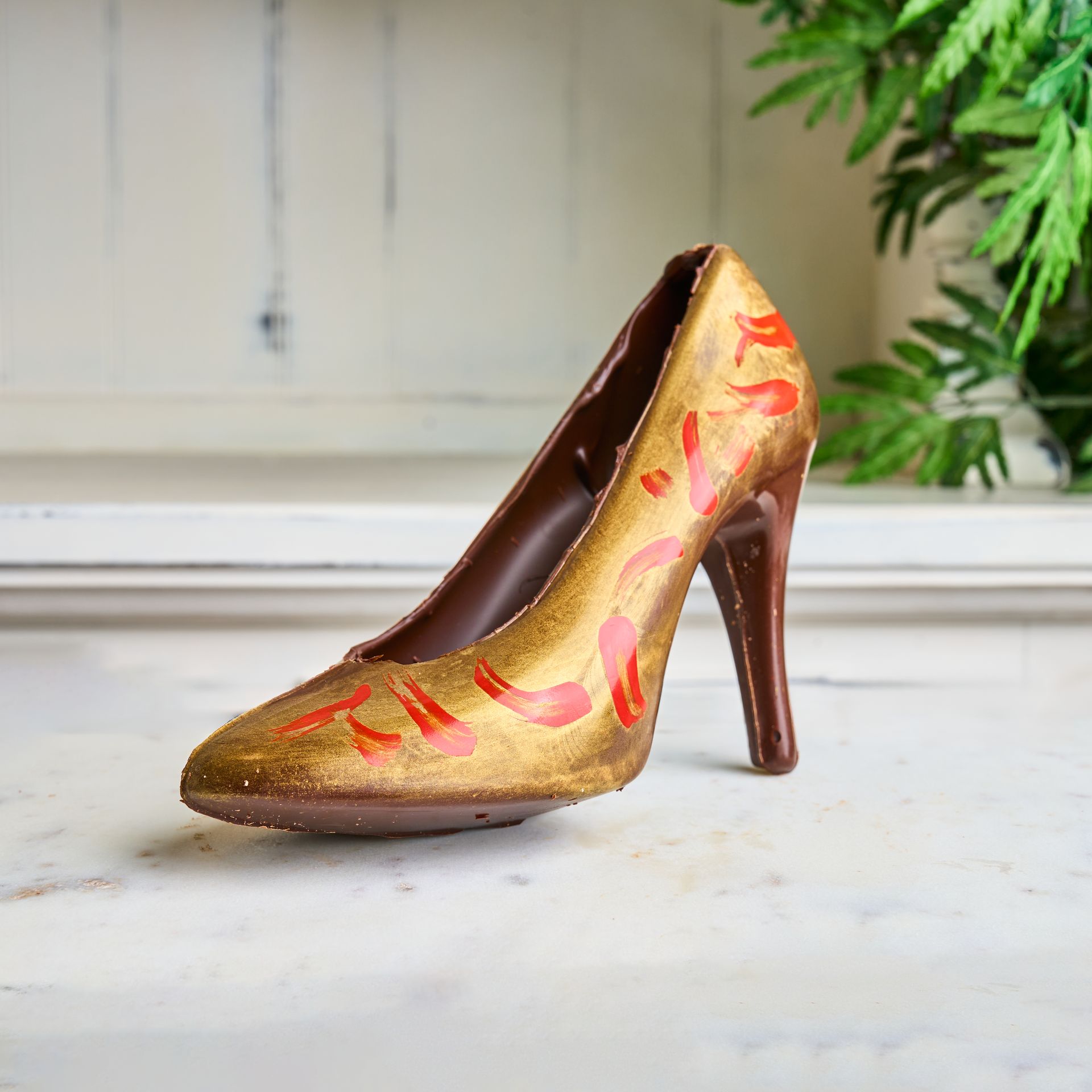 Chocolate high-heeled shoe, gold and red design, on white surface.