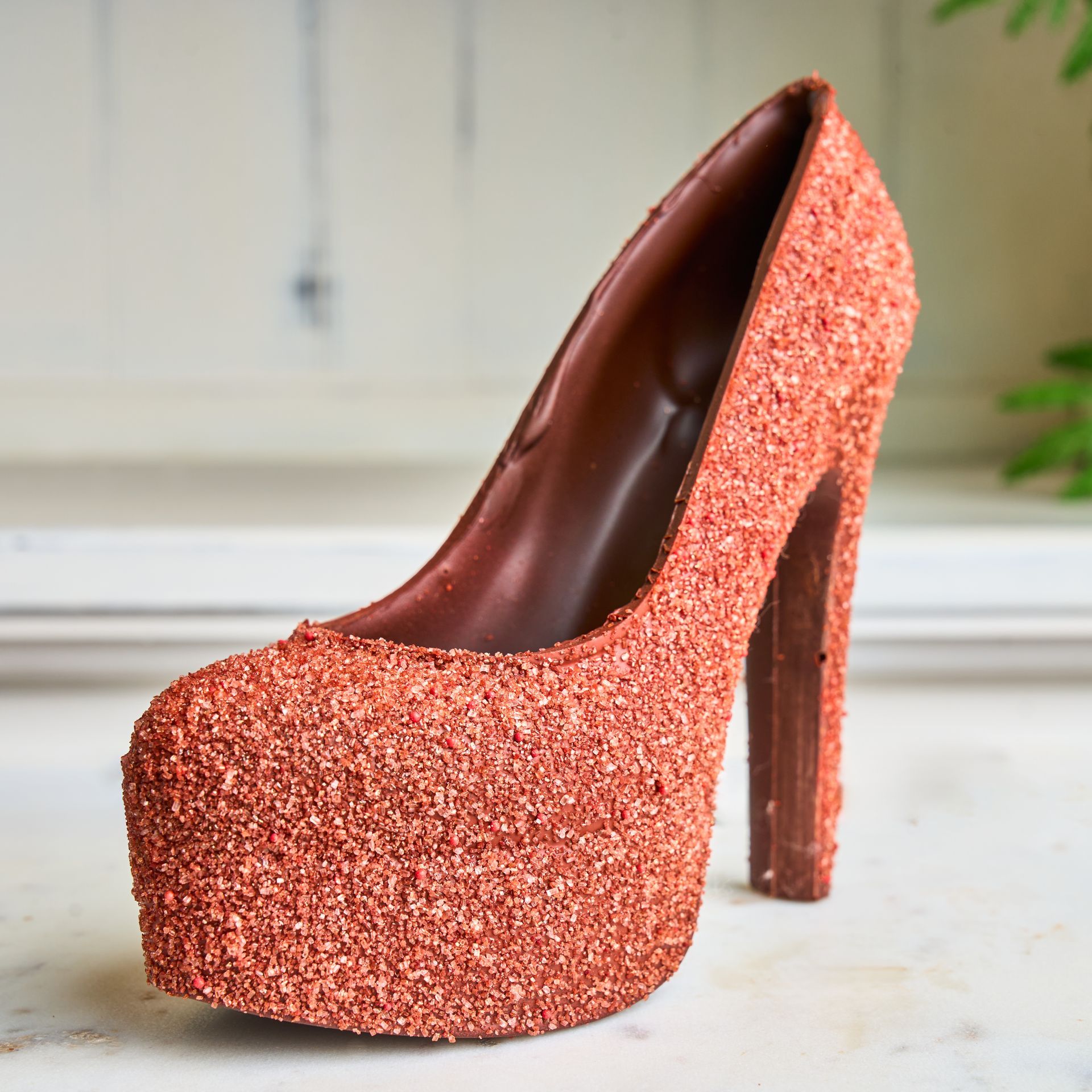 Chocolate high-heeled shoe, covered in sparkling copper-colored glitter, sitting on a white surface.