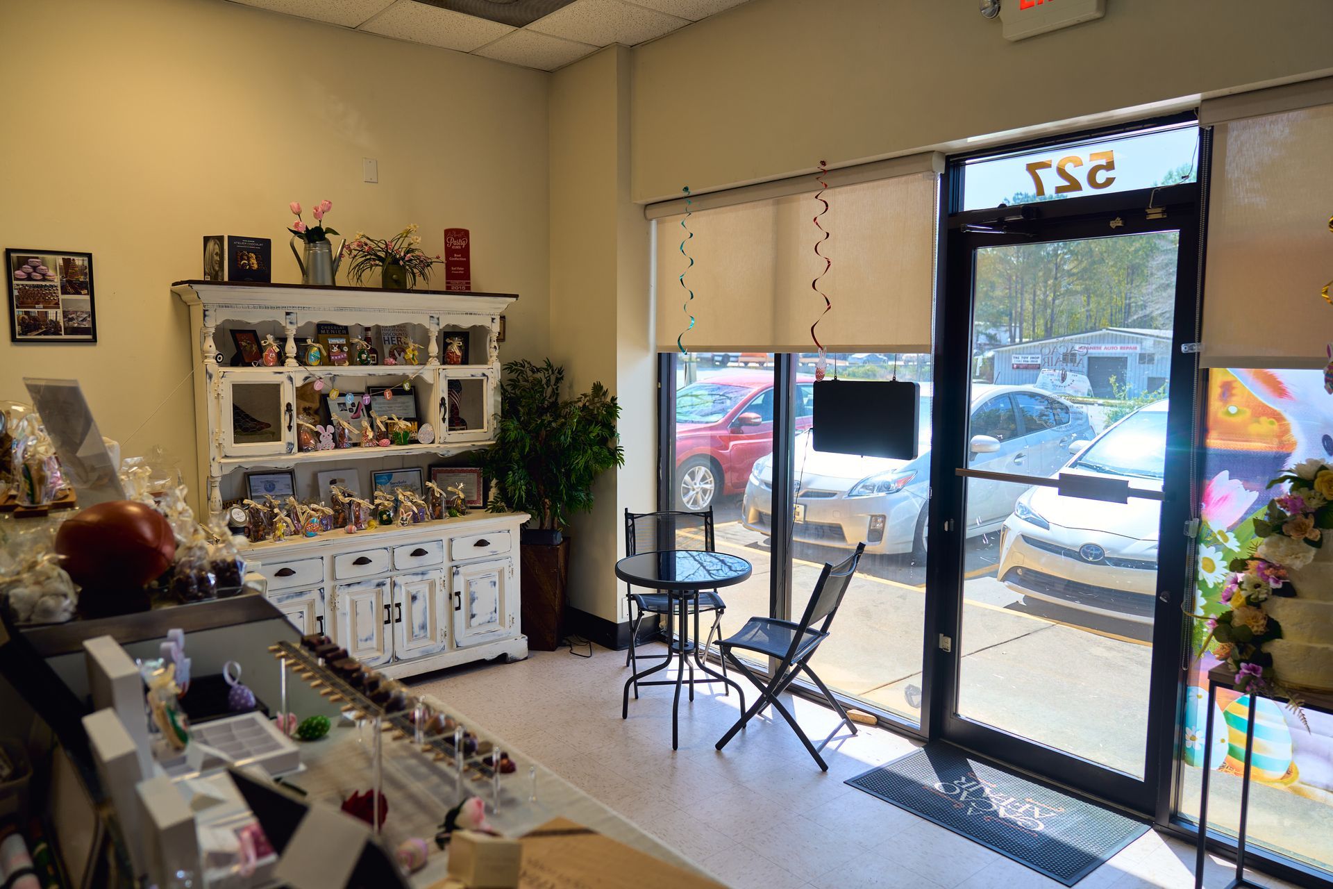 Interior of a shop with display shelves, a small table and chairs, and a window overlooking a parking lot with cars.