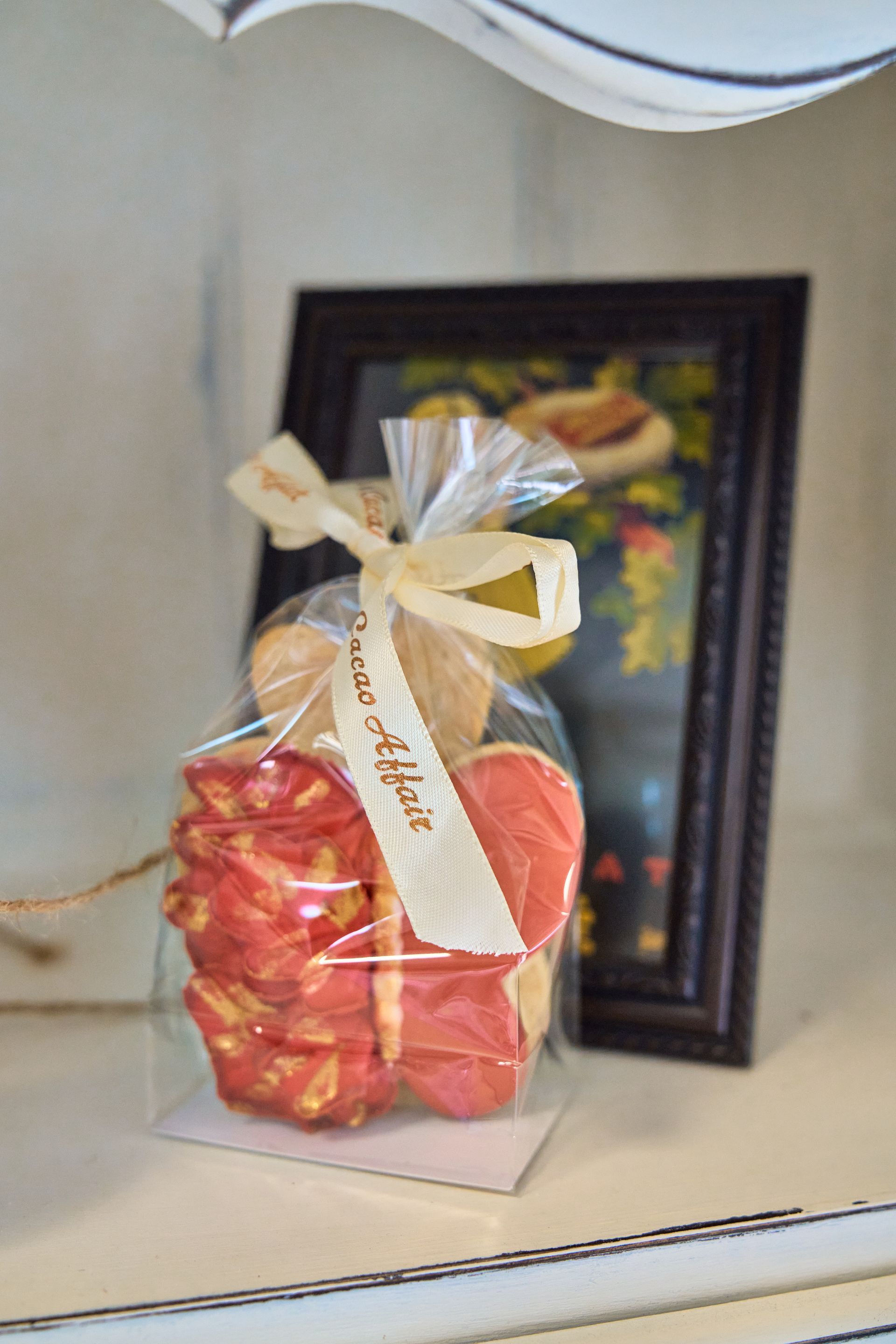 Cookies in a clear bag tied with a cream ribbon, in front of a framed painting on a shelf.