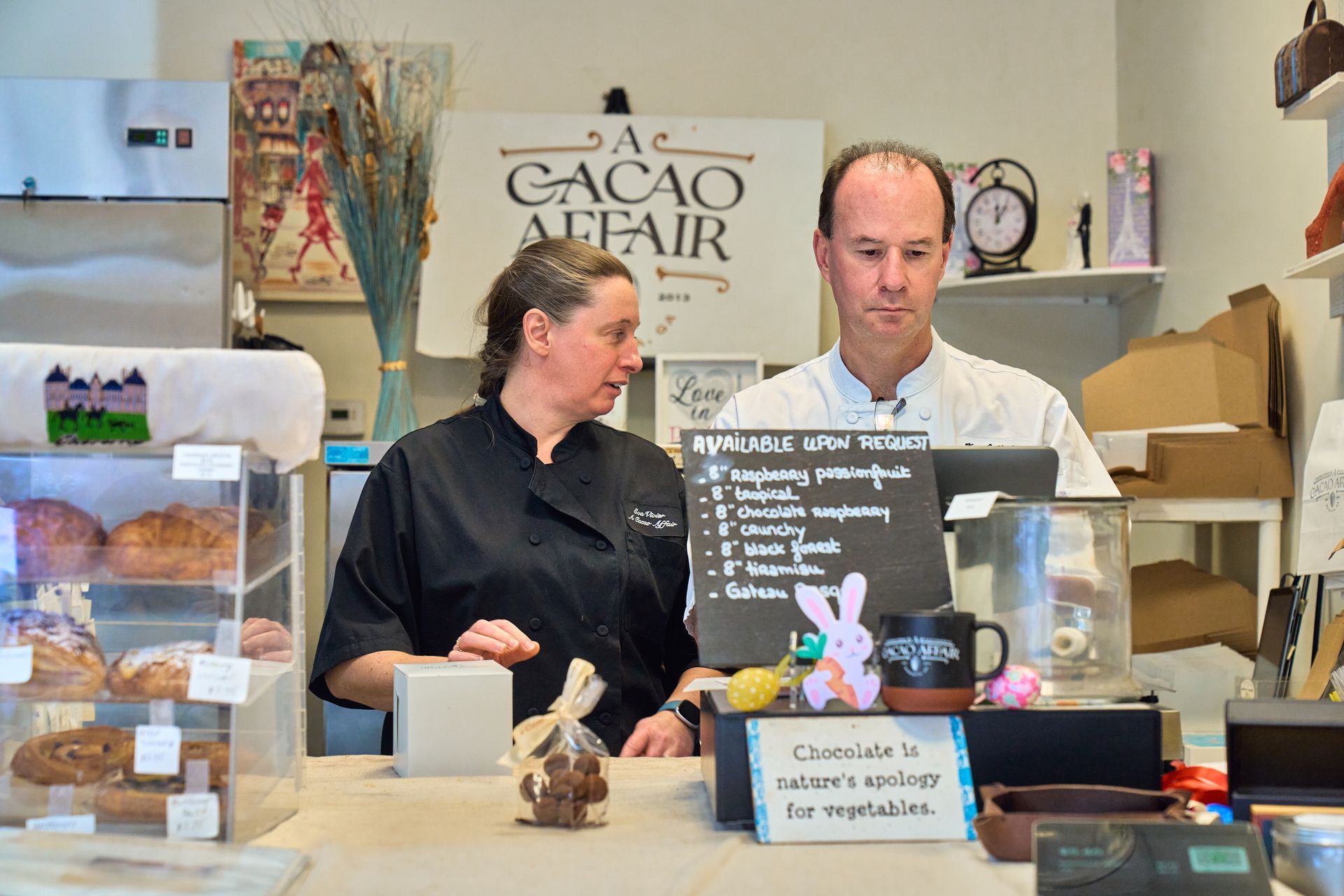 Two people at a bakery counter. A woman in black points; a man looks on. Display of treats and menu.