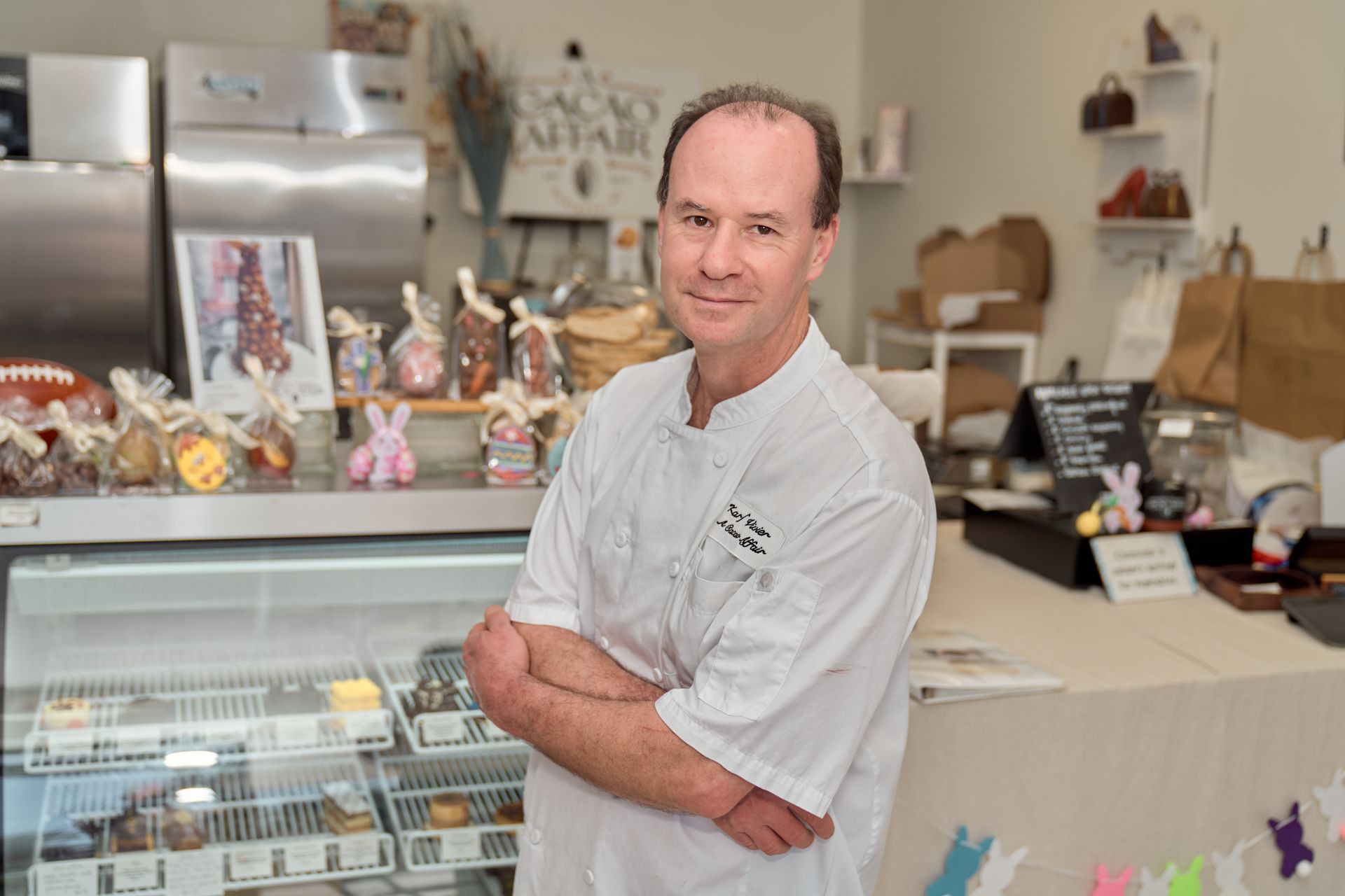 Man in white chef's coat, arms crossed, smiling in a chocolate shop. Display cases and treats in background.