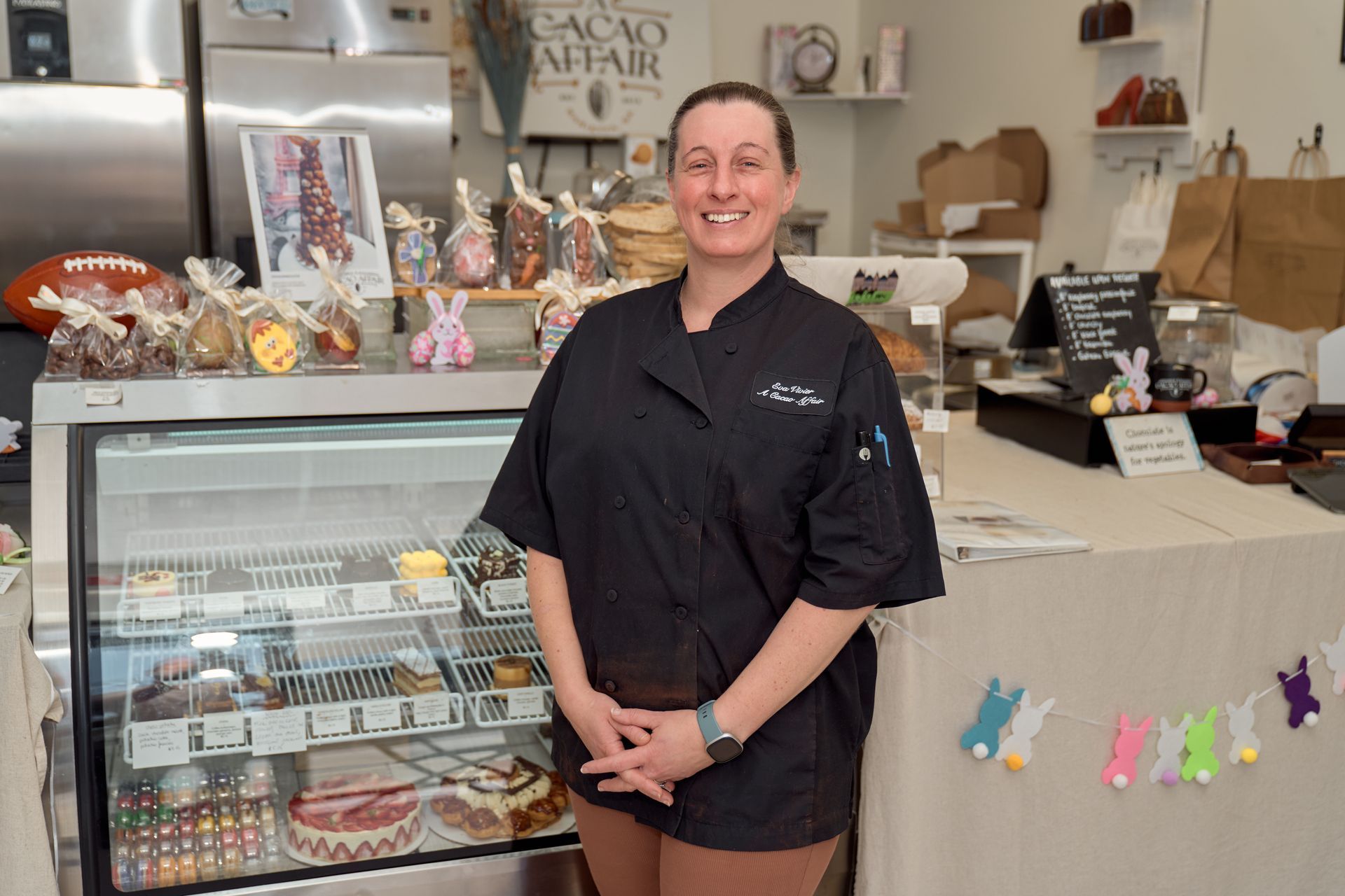 Woman in black chef coat stands in front of a glass display case filled with chocolates and candies.