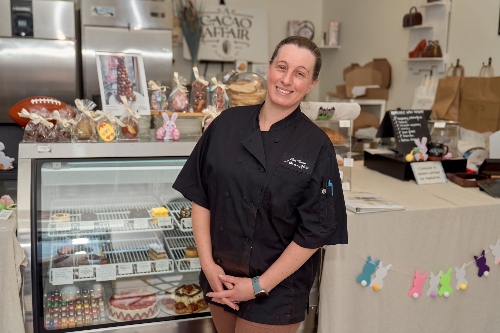 Woman in black chef's coat stands in chocolate shop, smiling. Baked goods, display case, and Easter decorations are visible.
