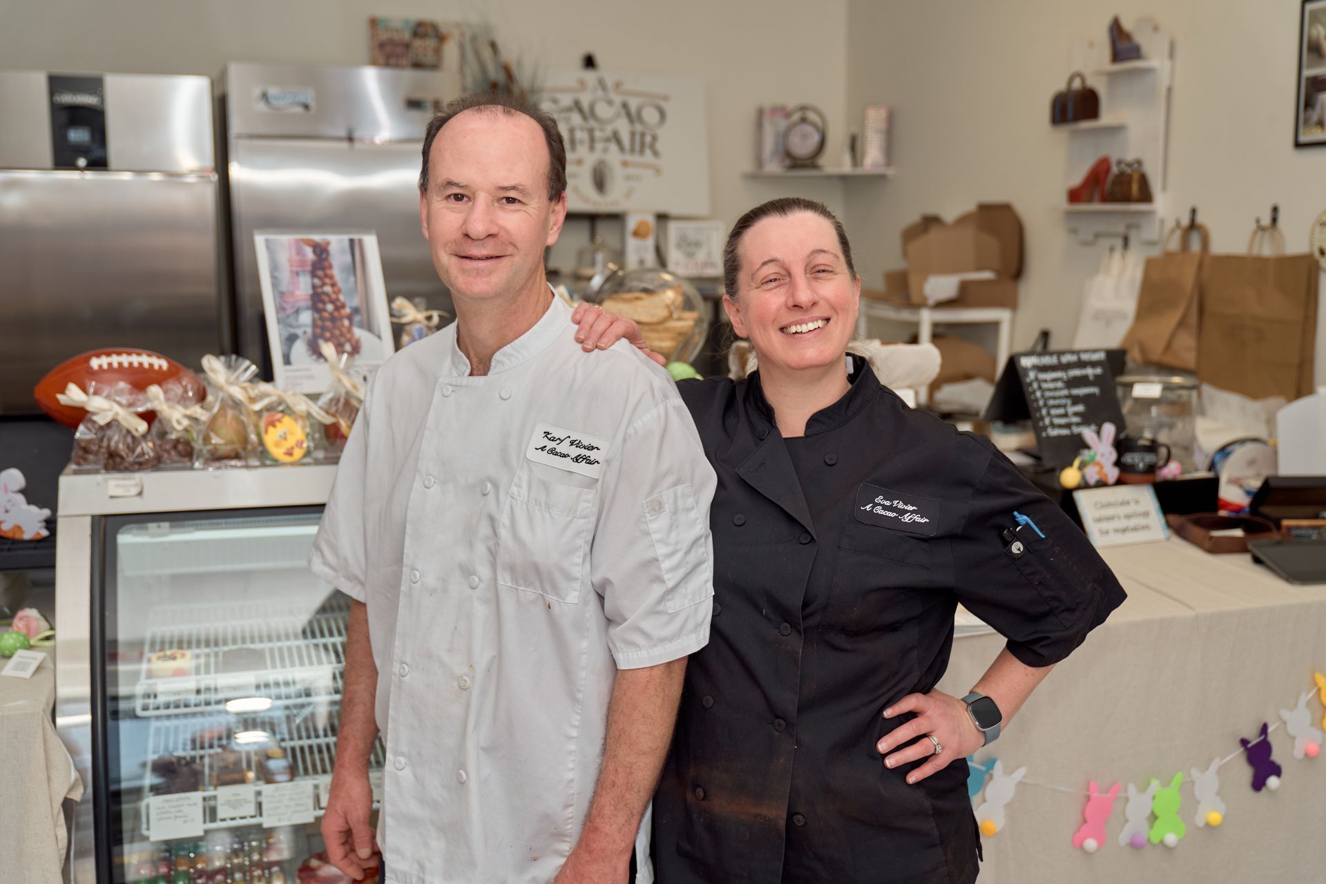 Two people in chef coats stand in a chocolate shop.