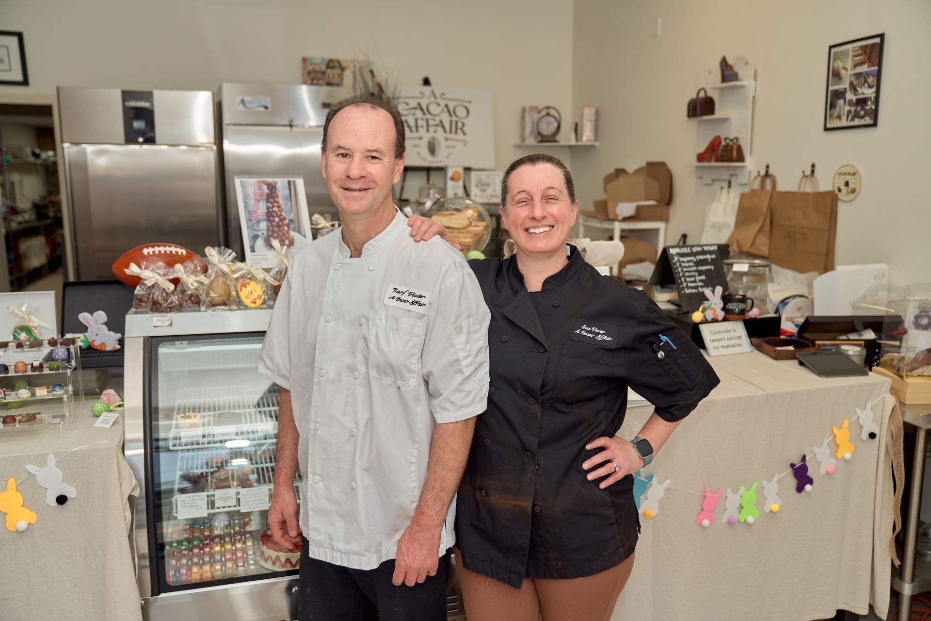 Two people in chef coats pose in a bakery. A display case and baked goods are visible.