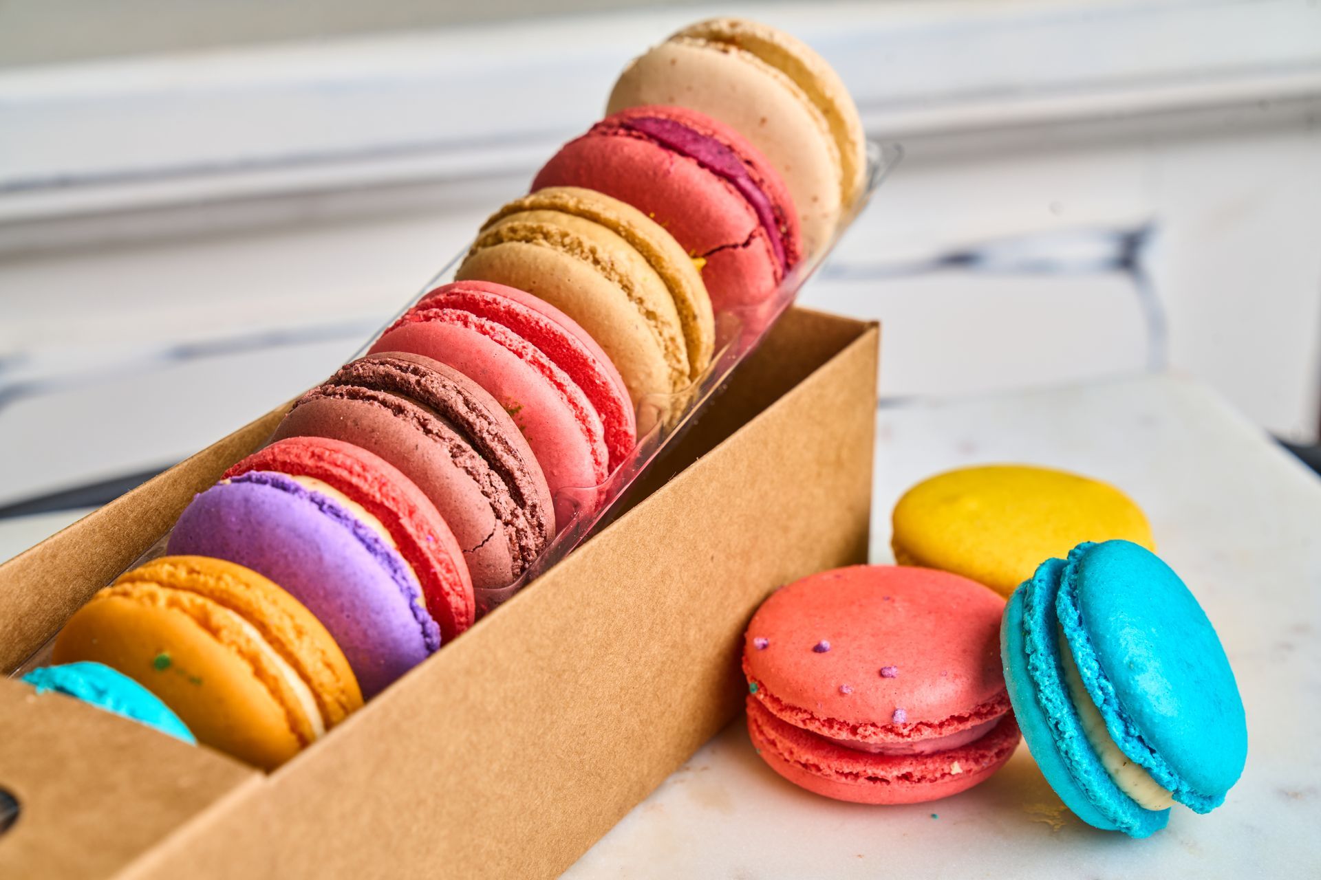 Colorful macarons in a cardboard box, with a few macarons outside the box, on a white surface.