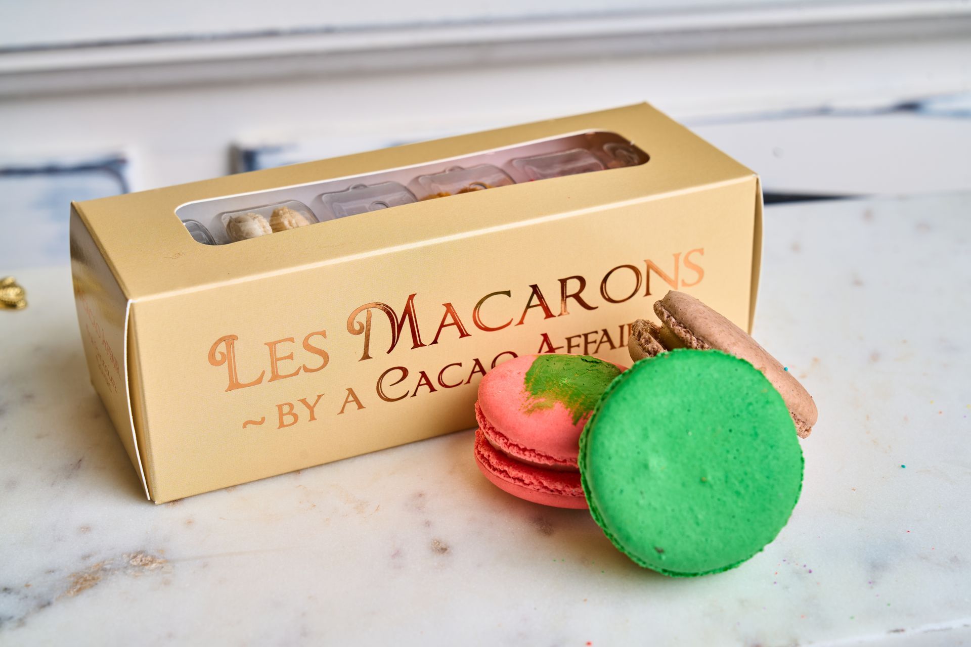 Box of macarons with a clear window; gold box, colorful cookies.