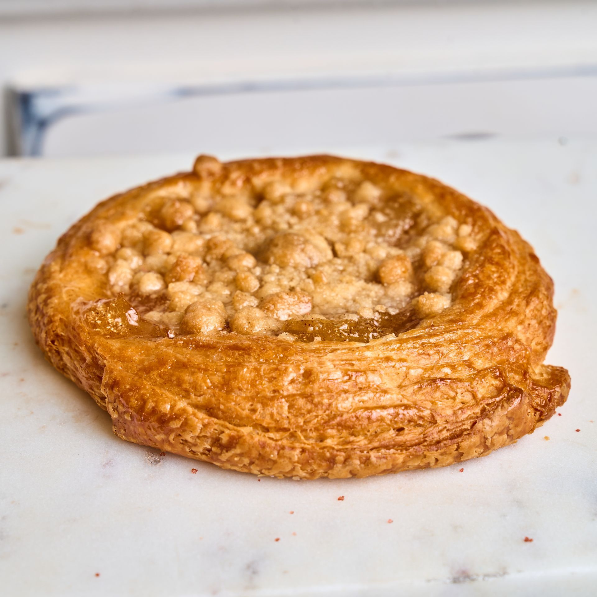 Round pastry with golden, flaky crust and crumble topping on a marble surface.