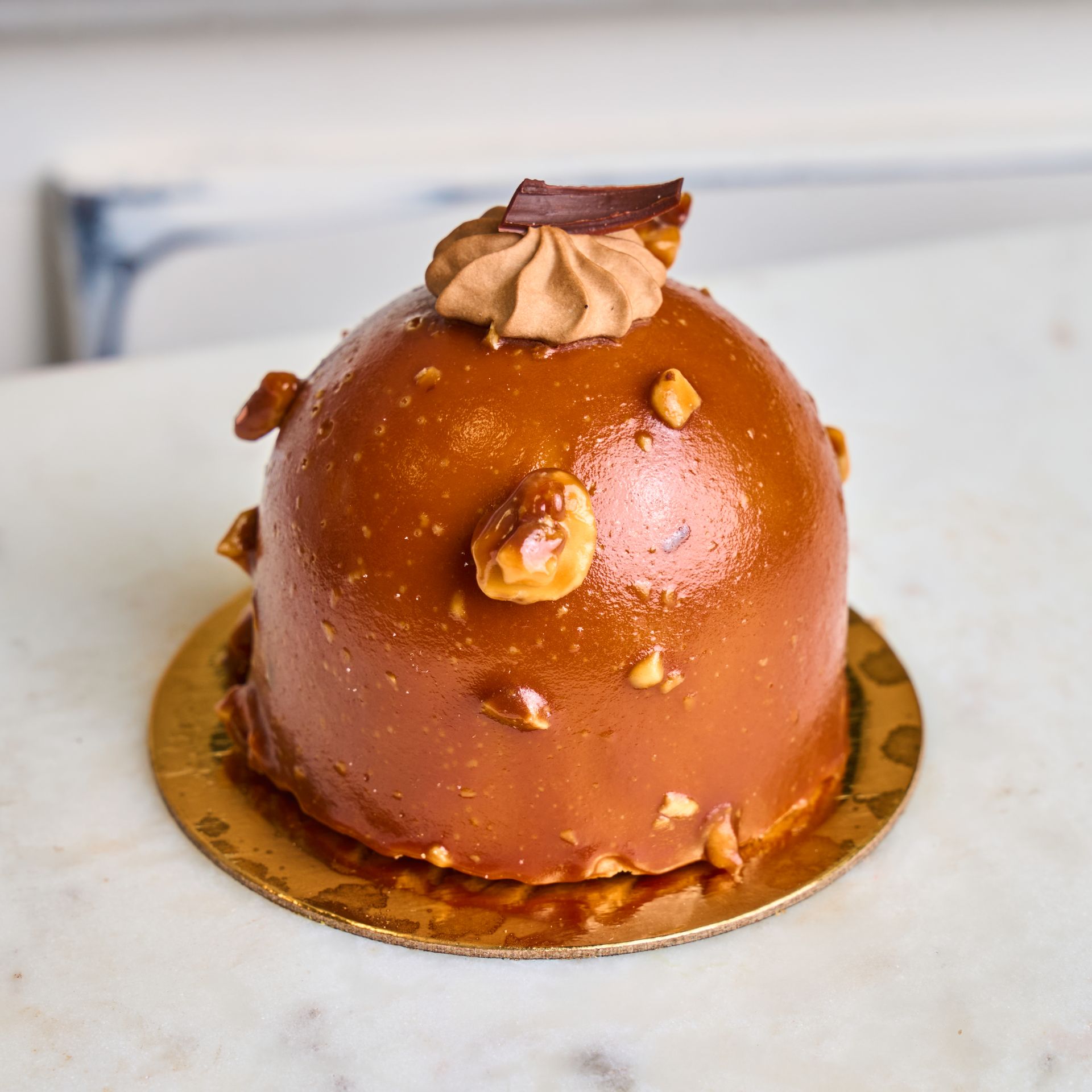 Dome-shaped caramel cake with nuts on a golden base, topped with frosting and a chocolate piece.