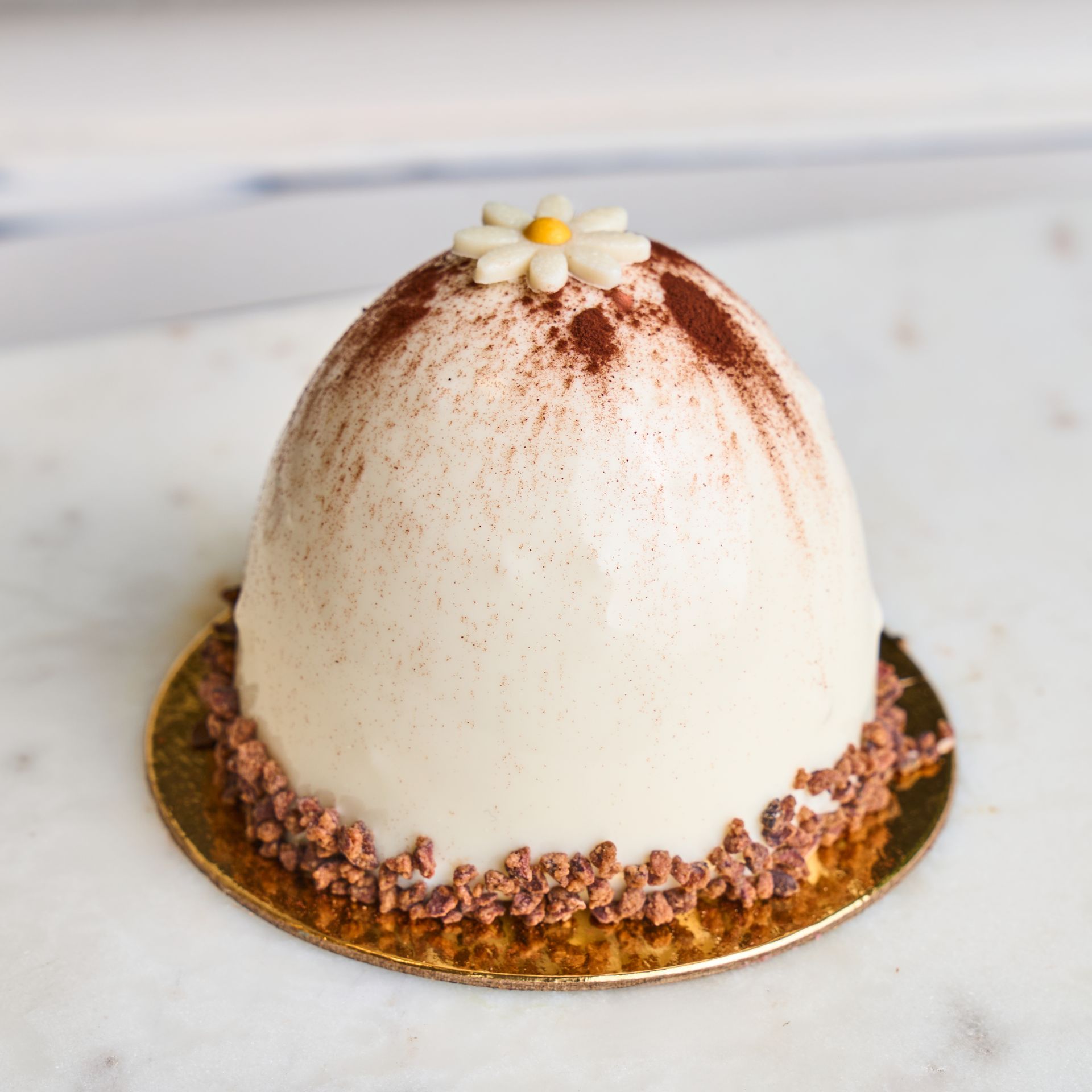 Dome-shaped white dessert dusted with cocoa, a small daisy on top, and chocolate sprinkles on a gold base.