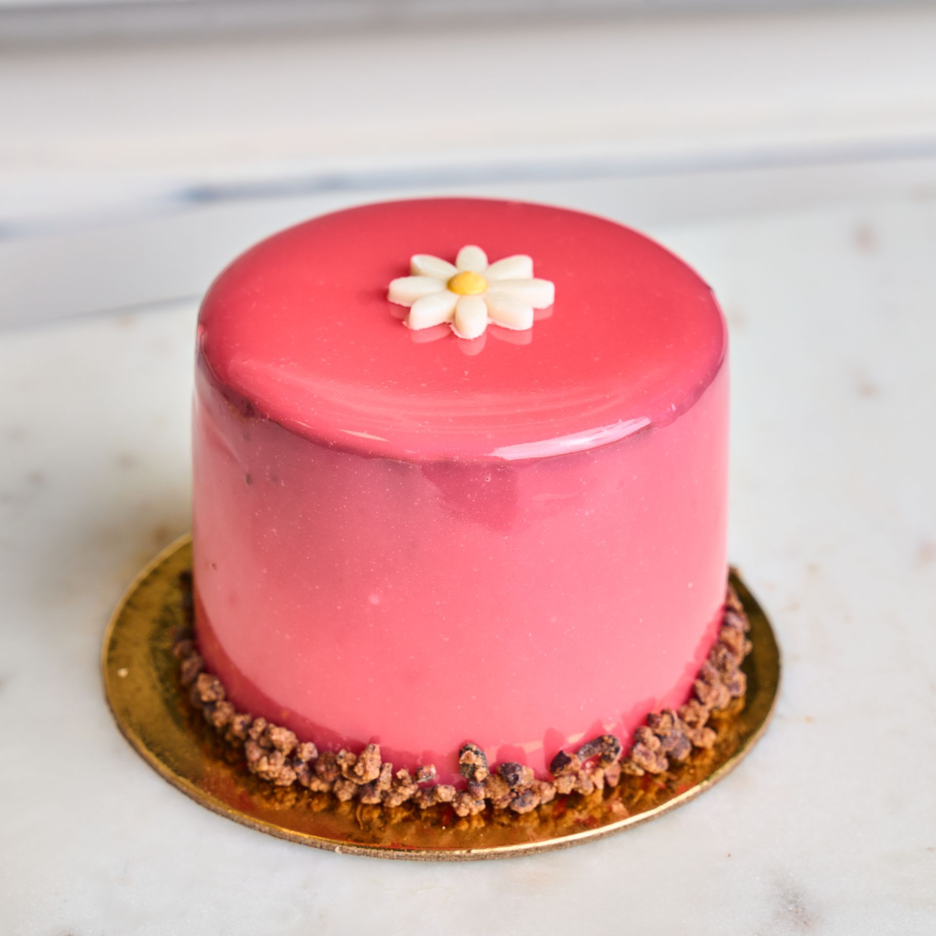 Pink glazed cake with daisy, on gold coaster. Chocolate crumble border.