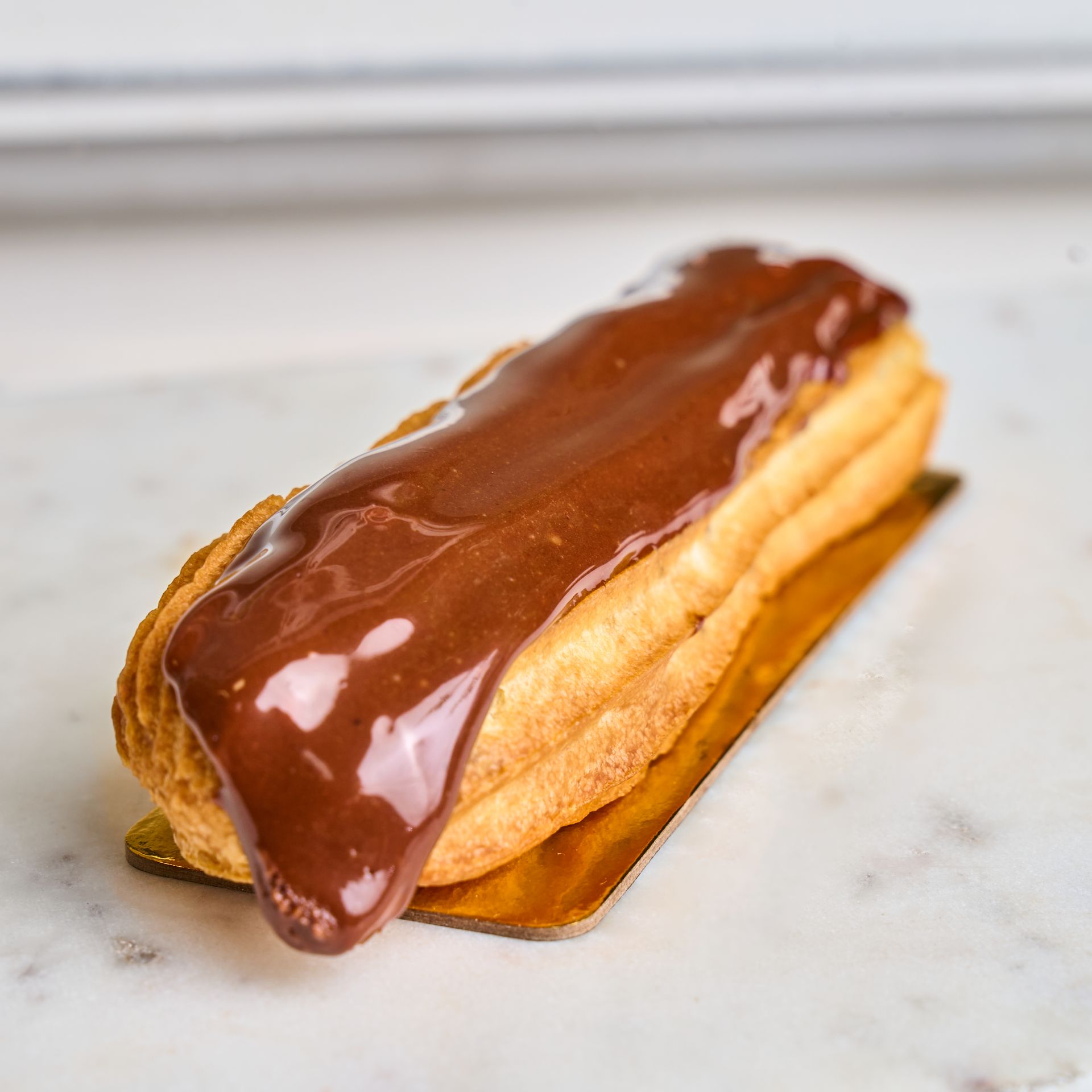Chocolate eclair on a gold paper tray, resting on a white marble surface.