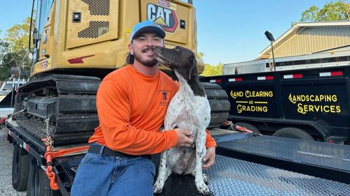 Man in orange shirt and dog on a trailer in front of construction equipment; dog is licking man’s face.