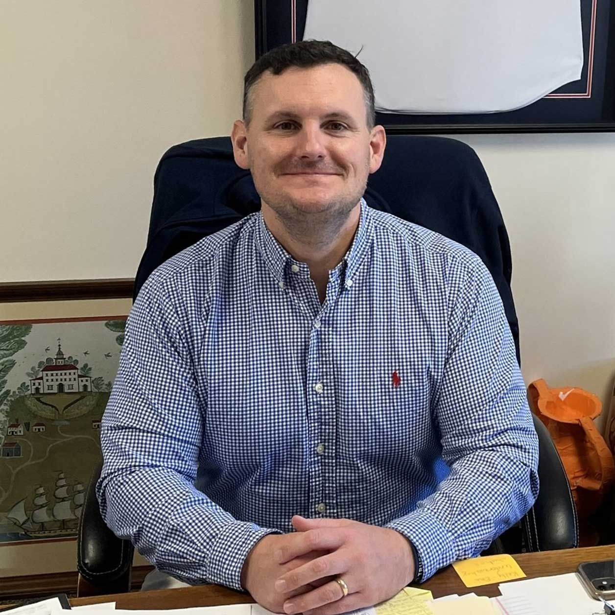 Man seated at an office desk in a blue checkered shirt, smiling at the camera.