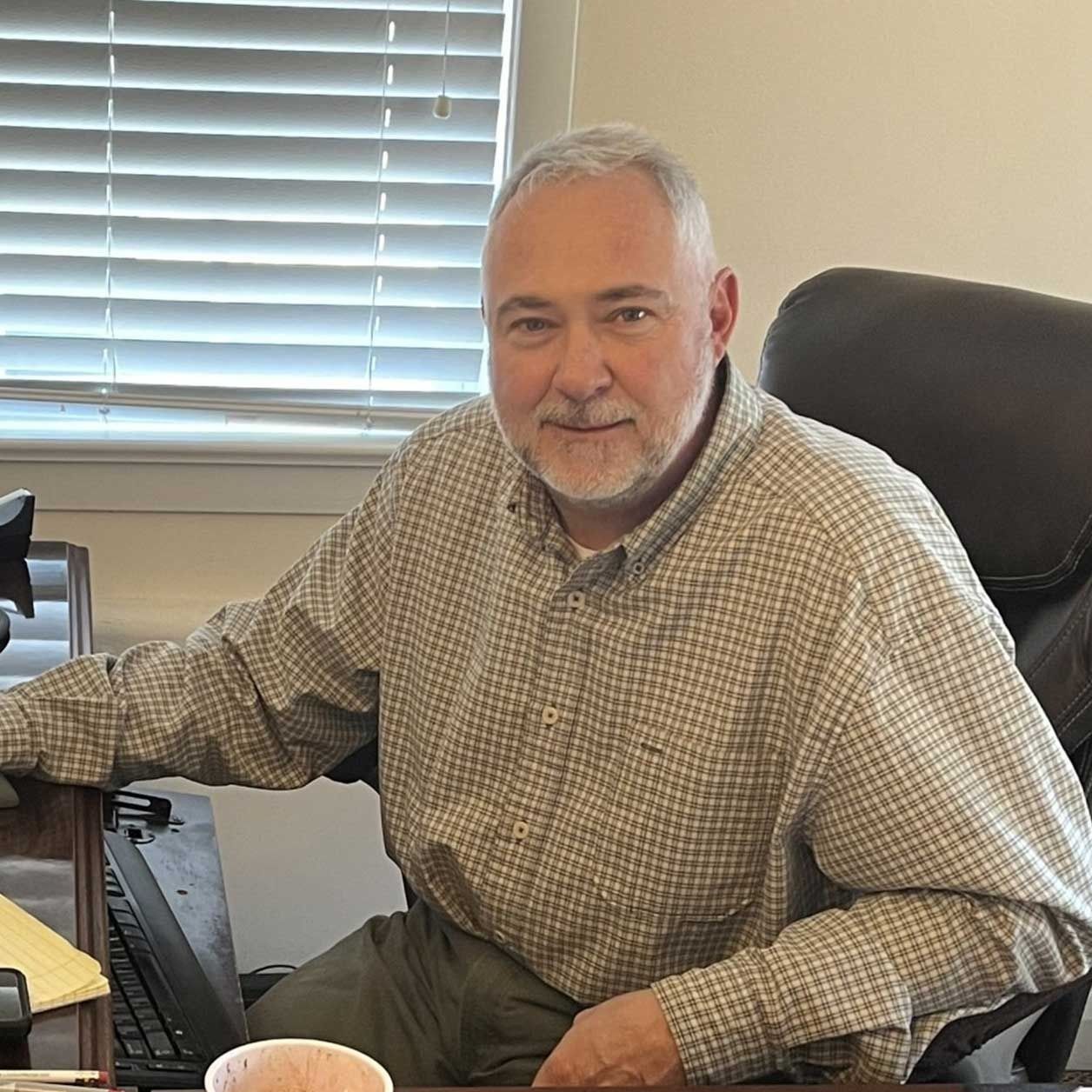 Man seated at a desk in an office, wearing a plaid shirt and looking at the camera.