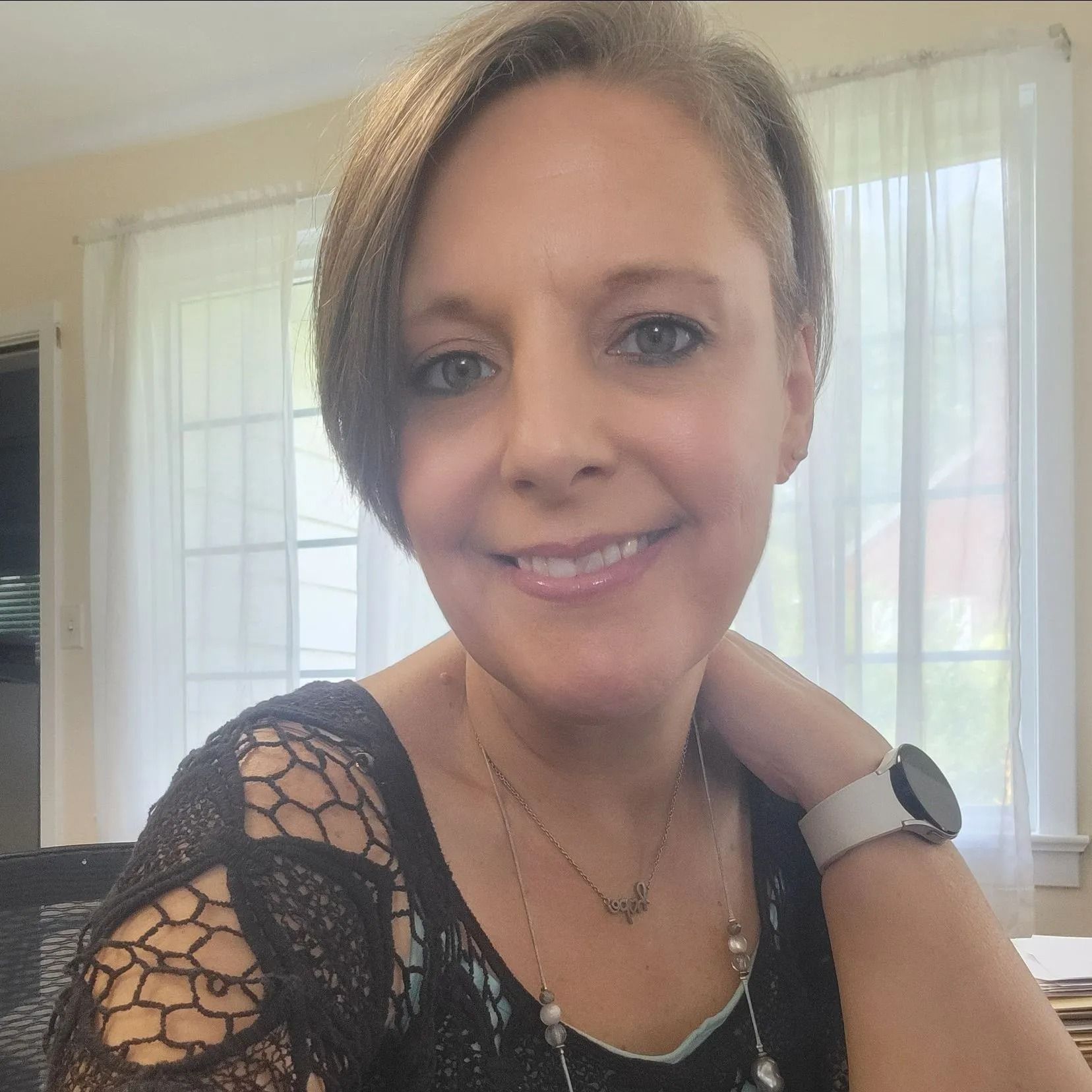 Smiling woman indoors by a bright window, wearing a black lace top and watch.