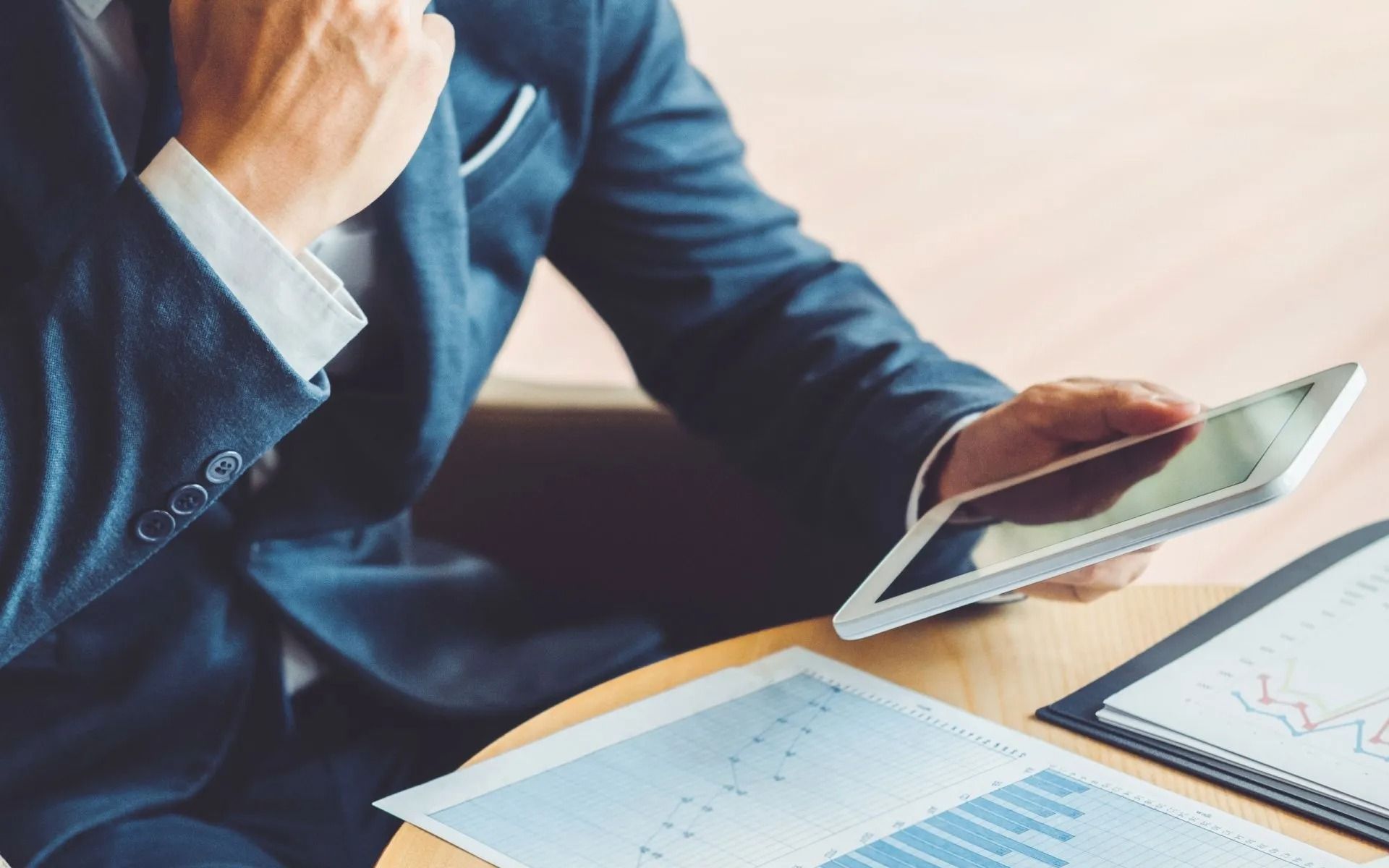 Businessperson in blue suit reviewing a tablet beside financial charts and documents on a desk