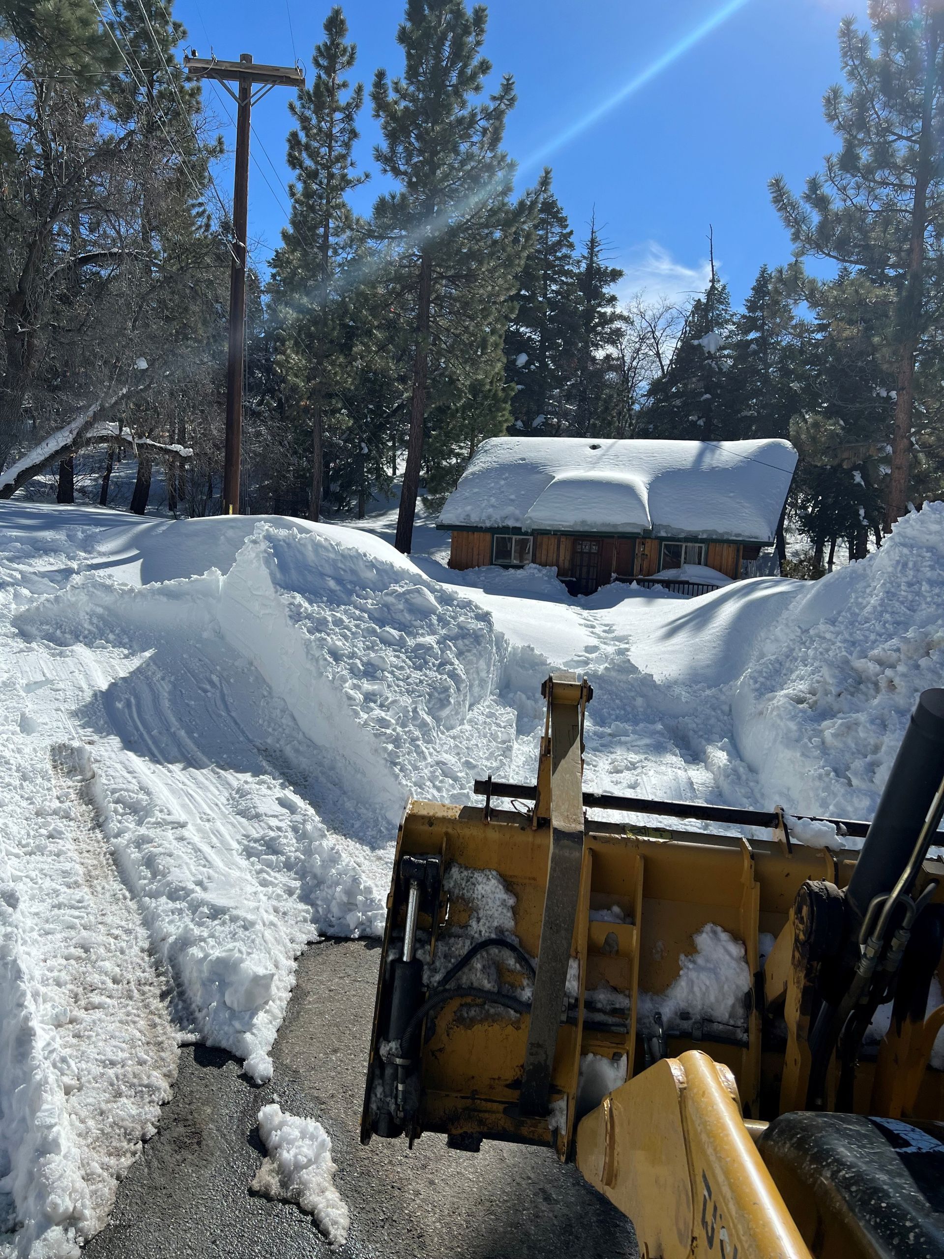 Snowplow clearing a snow-covered road near a cabin. Sunny day, bright sky, snow piled high.