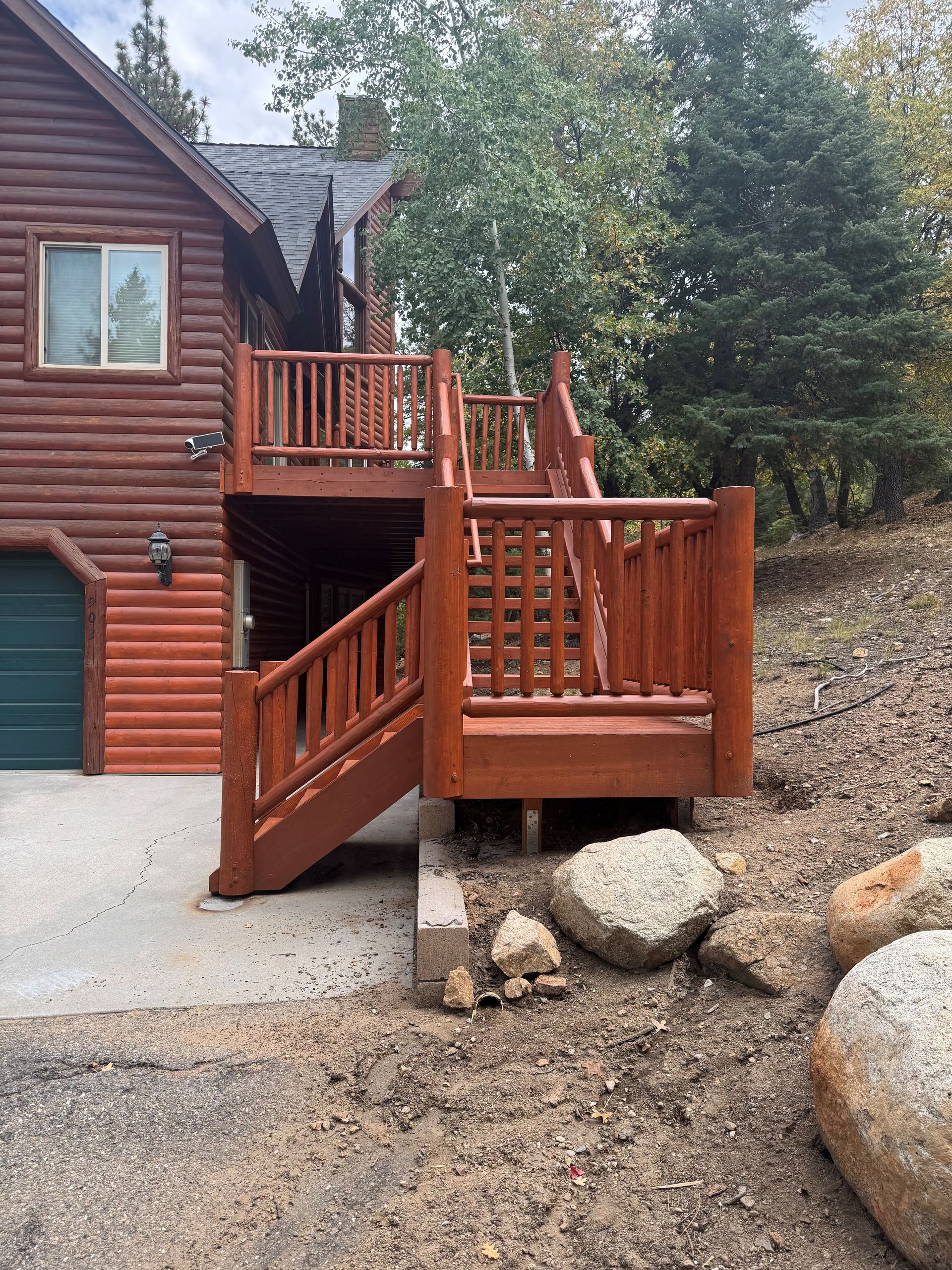 Wooden deck and stairs painted brown, leading up to a log cabin. The ground is gravel and has some boulders.