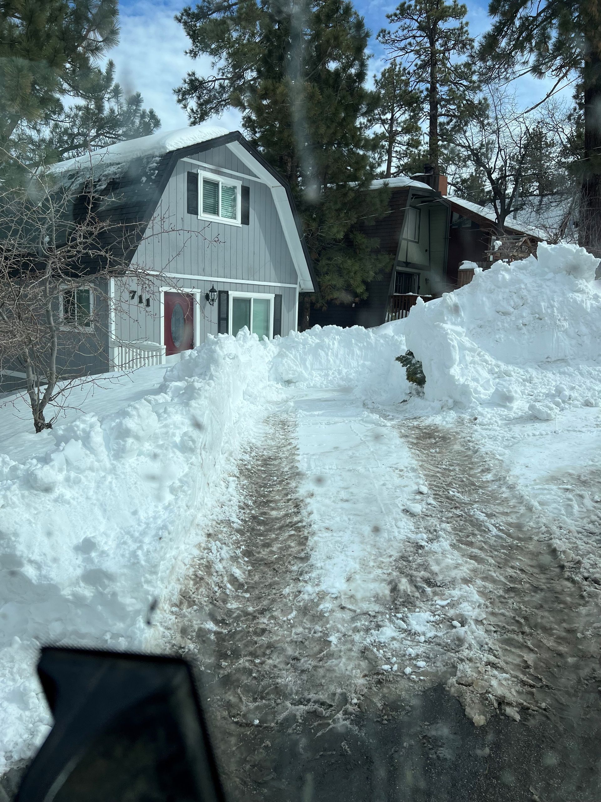 Snowy driveway leading to a gray house.  Large snowbanks flank the path.  Blue sky peeks through trees.