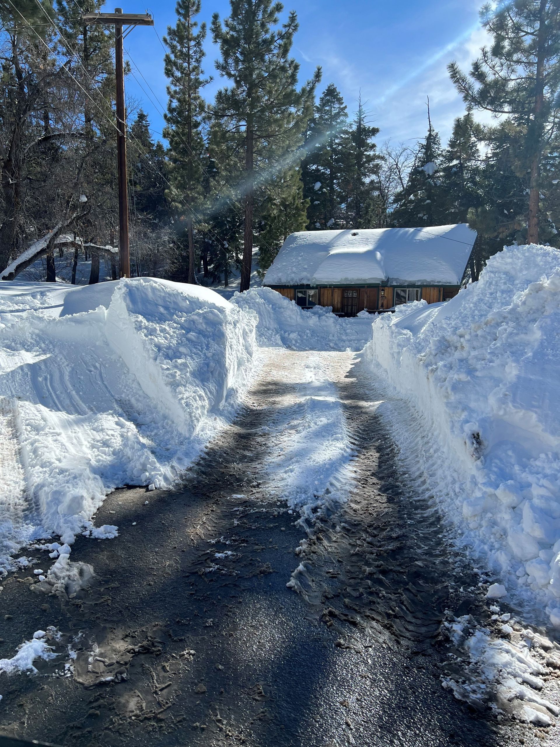 Snow-covered driveway leading to a wooden cabin. Tall snow banks line the path under a blue sky.