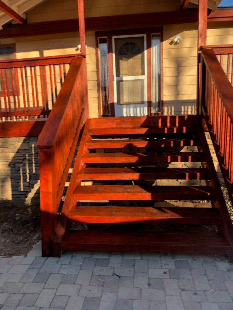 Wooden stairs and deck leading to a house entrance; red-stained wood, stone facade, and paved patio.