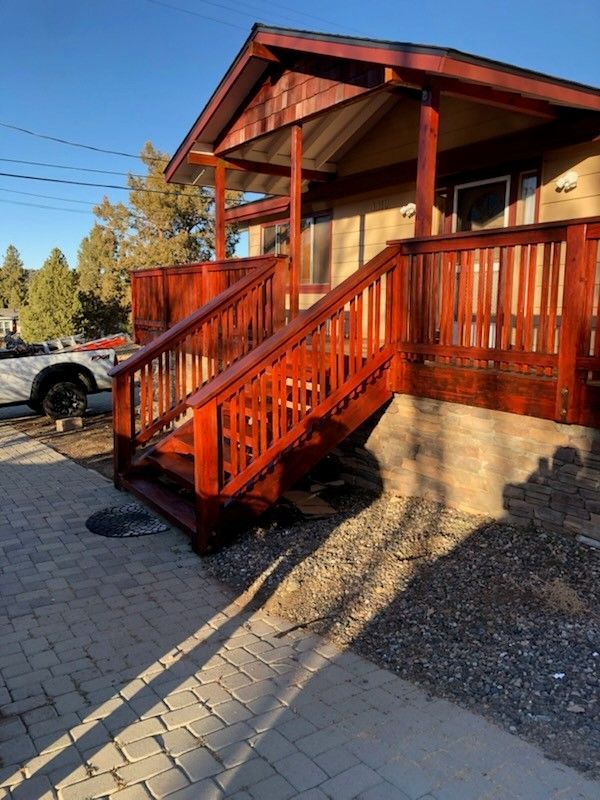 Wooden porch and staircase of a house, stained red-brown. Sunny day, paved walkway.