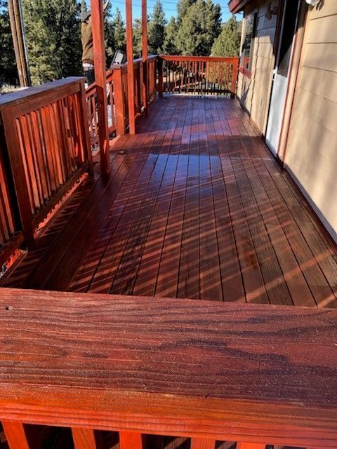 Wooden deck with red-stained railing and floor. Sunlight creates shadows across the wet surface.